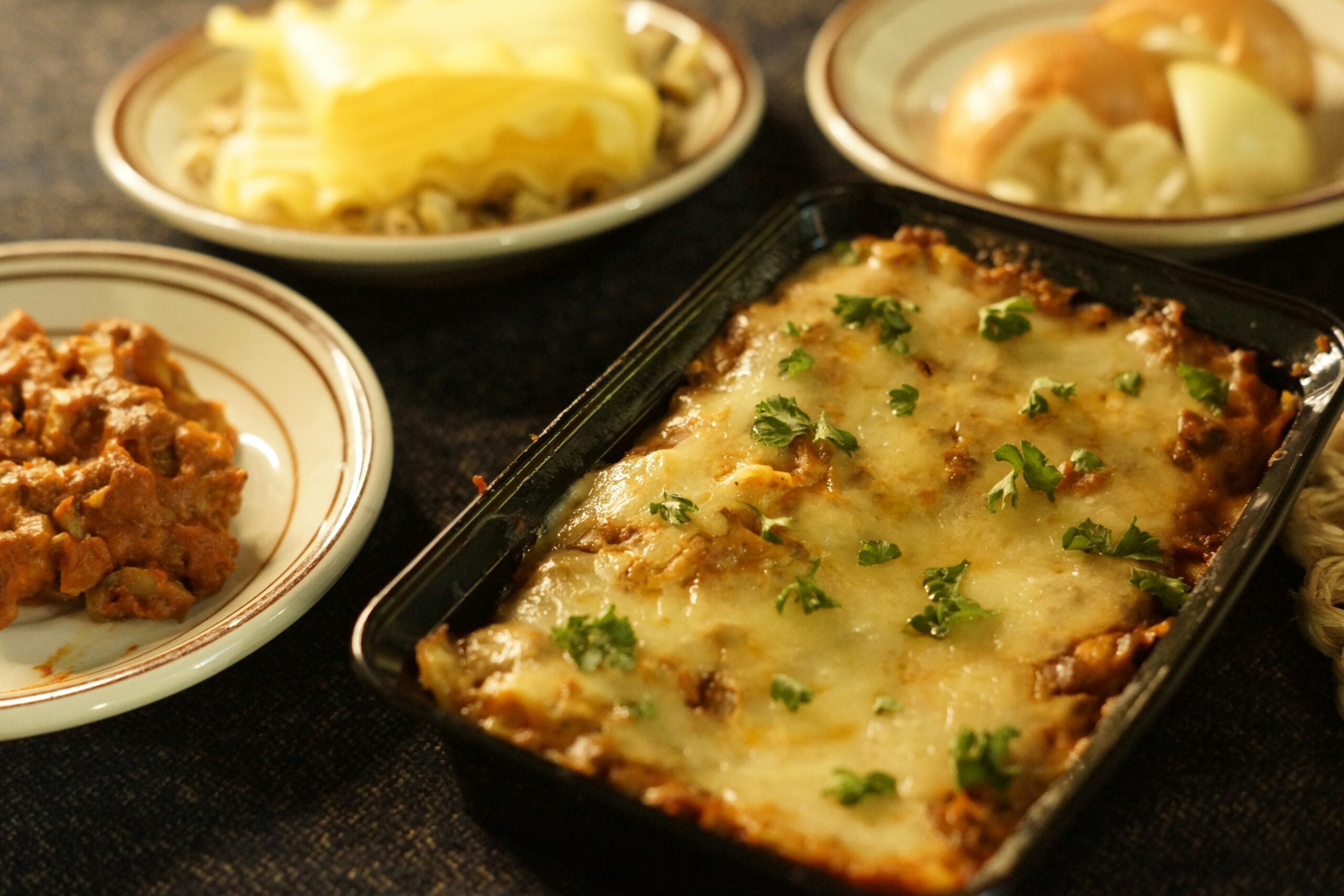 A rectangular dish of baked lasagna topped with melted cheese and parsley is surrounded by plates with pasta, bread rolls, and a serving of lasagna on a dark tablecloth.