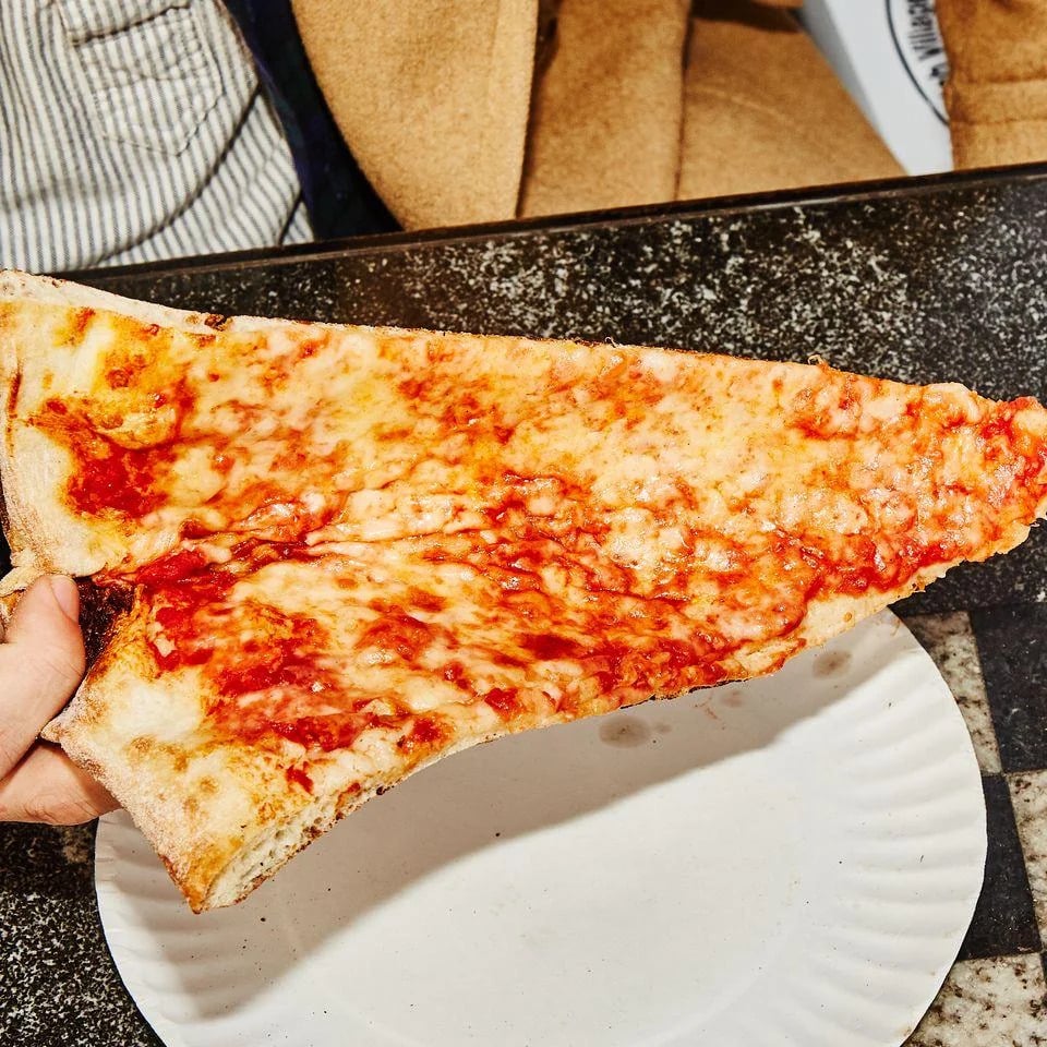 A hand holds a large, thin slice of cheese pizza with tomato sauce over a white paper plate on a dark countertop. A person's torso in a striped shirt and tan jacket is partially visible in the background.