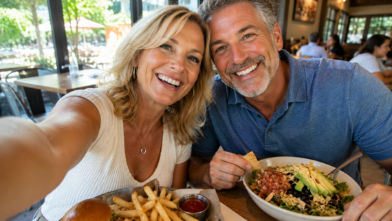 A smiling middle-aged couple takes a selfie at a sit-down restaurant table with a burger, fries, and salad in front of them. Bright daylight streams through large windows in the background.
