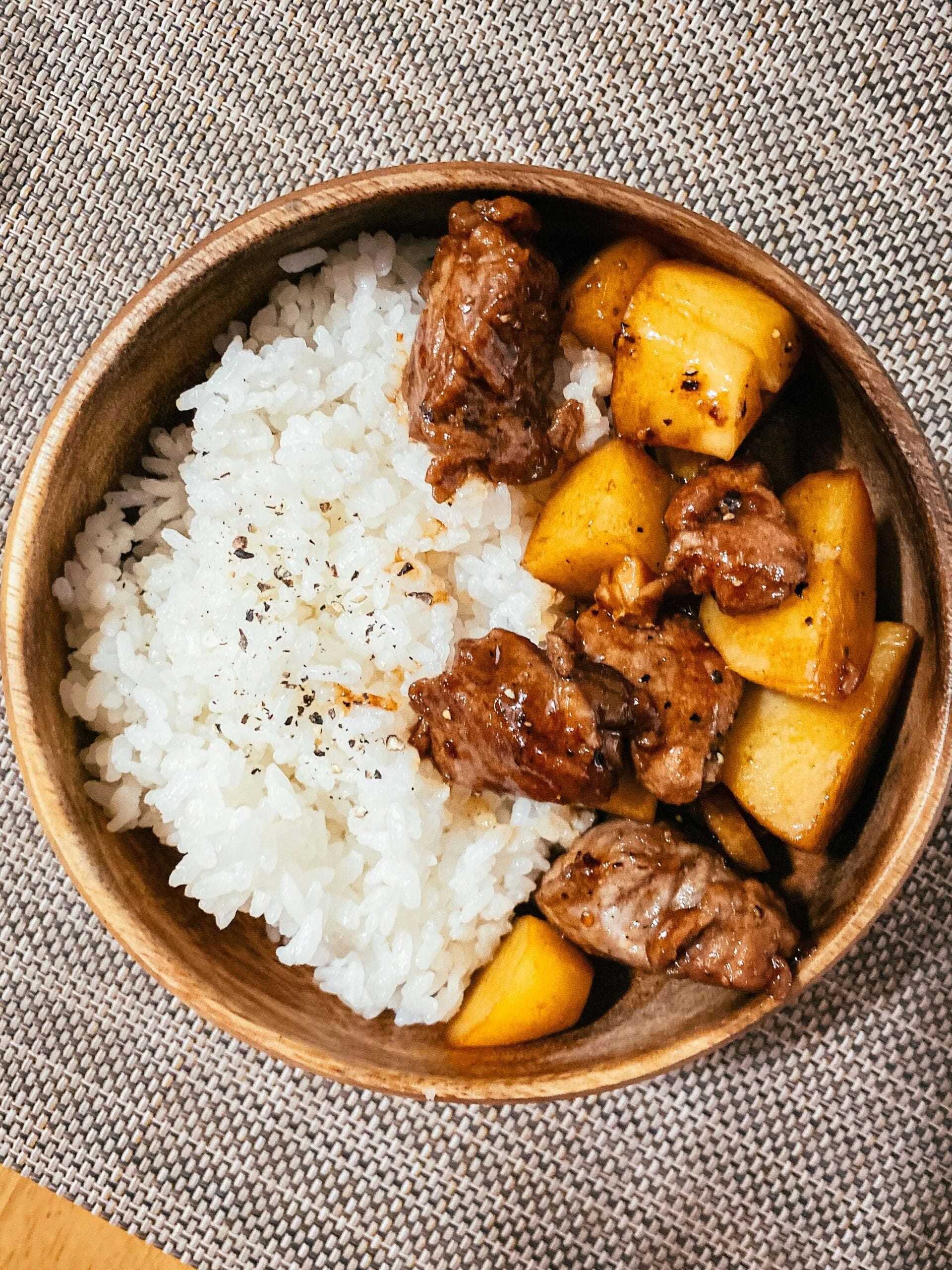 A wooden bowl containing white rice sprinkled with black pepper on one side and cooked meat with yellow potatoes on the other, placed on a woven placemat.
