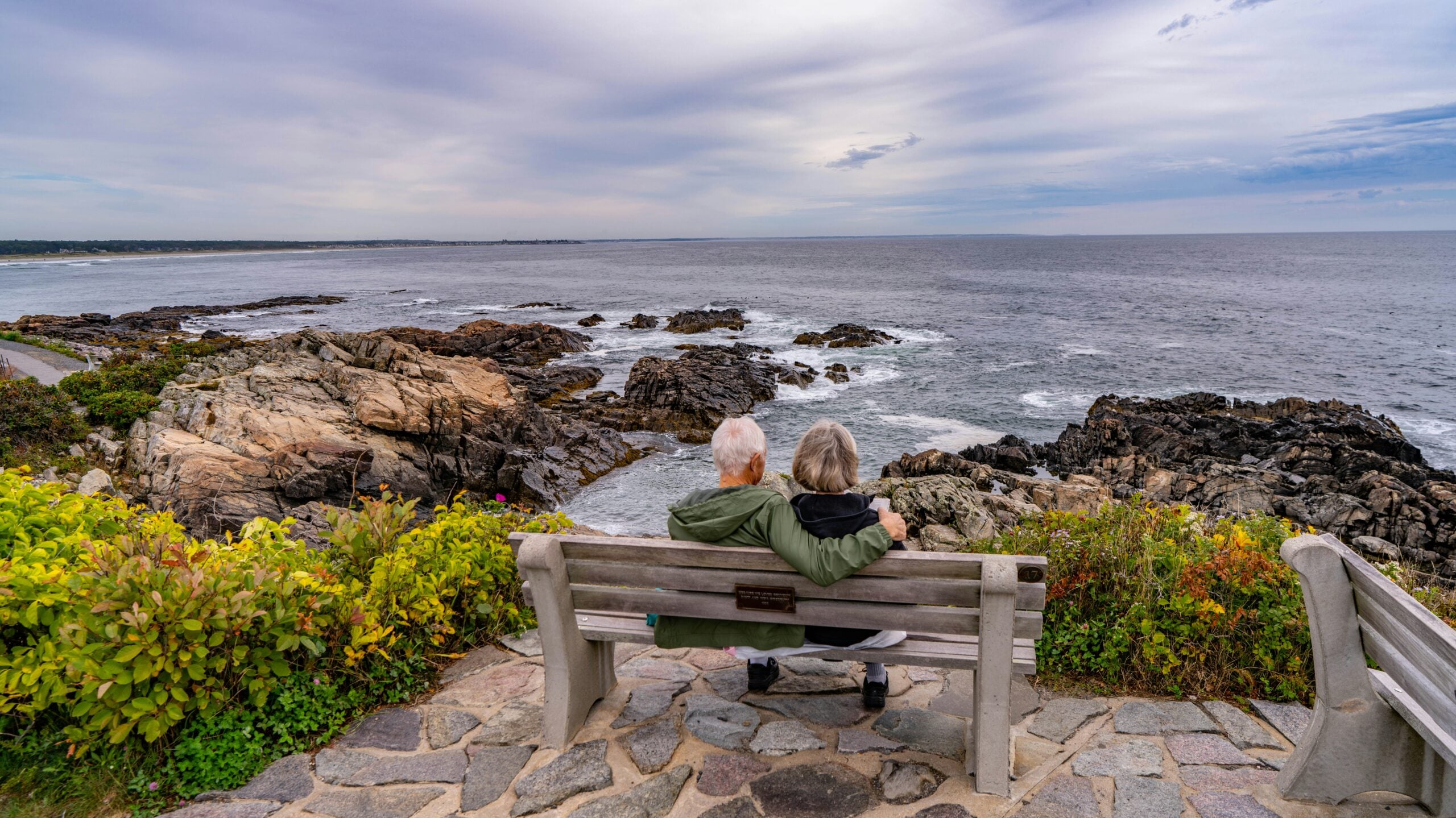 An elderly couple sits closely together on a wooden bench, overlooking a rocky shoreline and the ocean under a cloudy sky. Surrounded by lush greenery, they enjoy a peaceful moment, reflecting on Cost of Living Relief in this scenic setting.