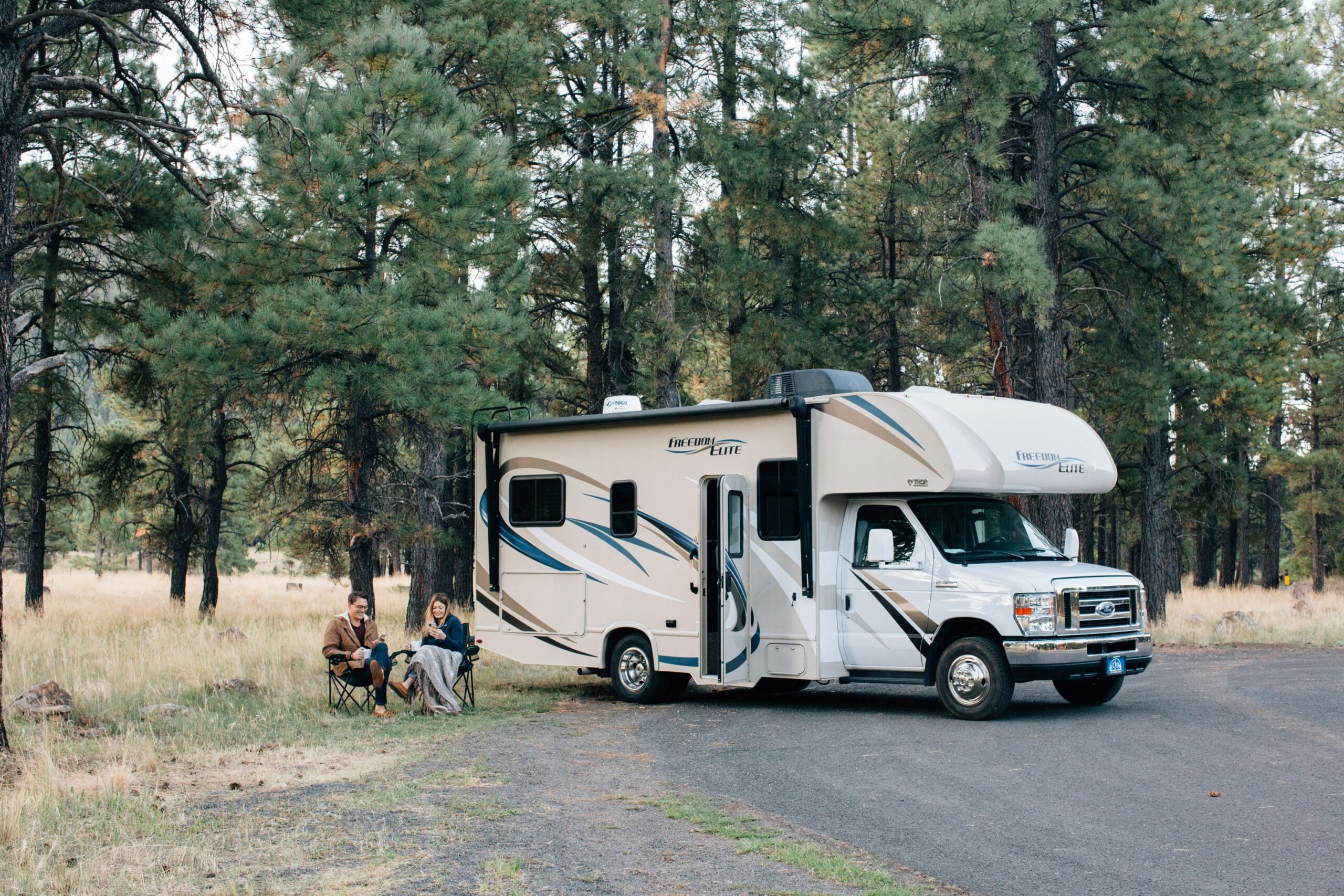 Two people sit on camping chairs outside a parked RV in a forest with tall pine trees. They appear to be relaxing, with the RV on a paved area surrounded by grass and trees.