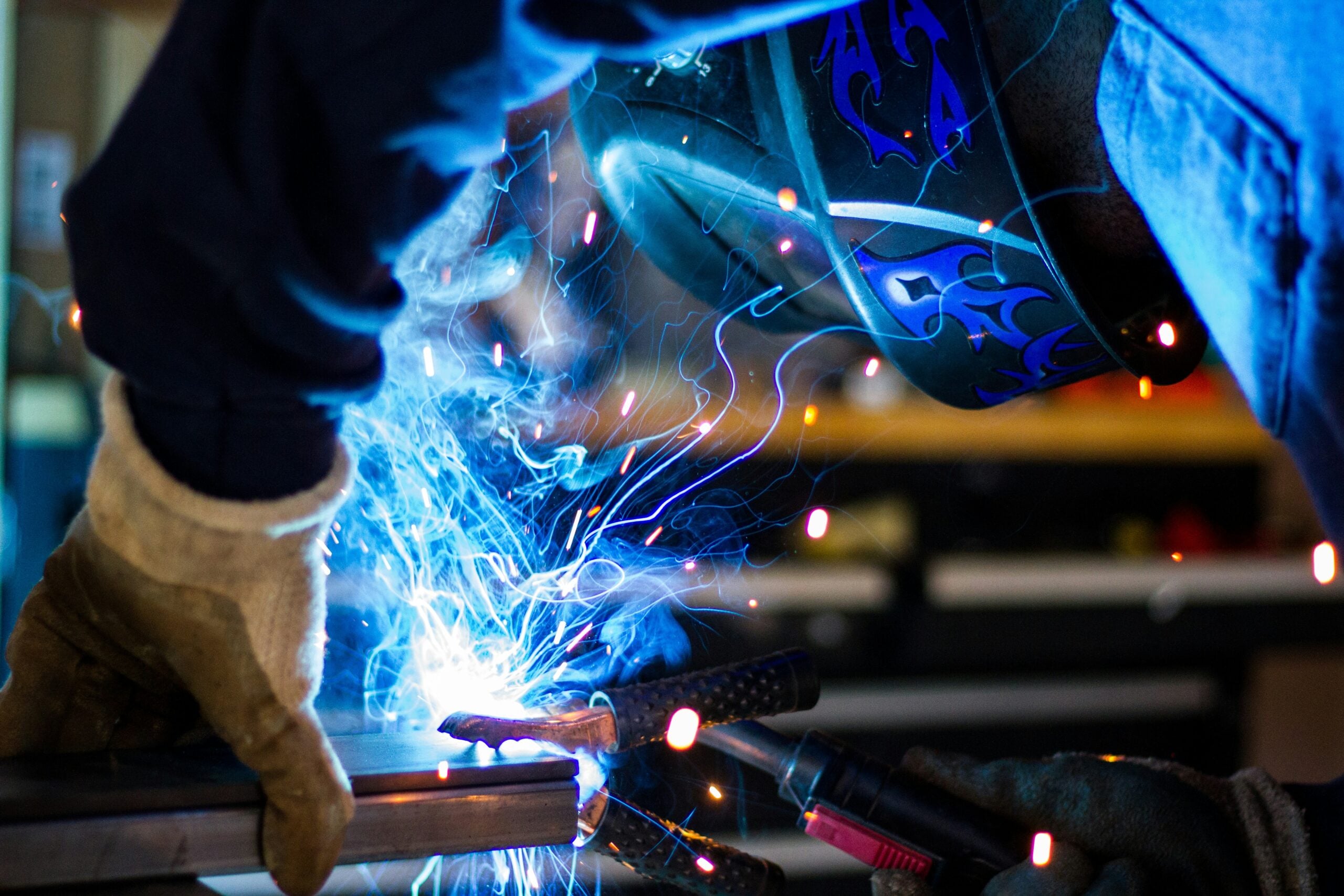 A person wearing a protective welding helmet and gloves welds metal, with bright blue sparks and smoke illuminating the scene.
