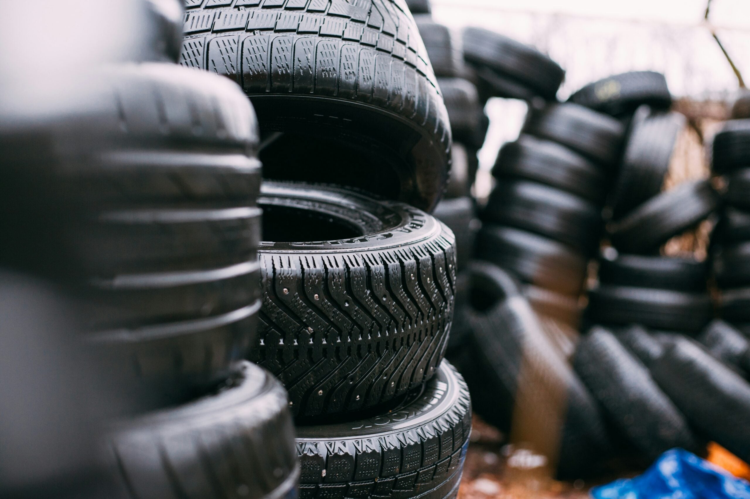 Stacks of black car tires, some neatly piled in the foreground and others scattered in the background, outdoors in a tire yard or storage area.