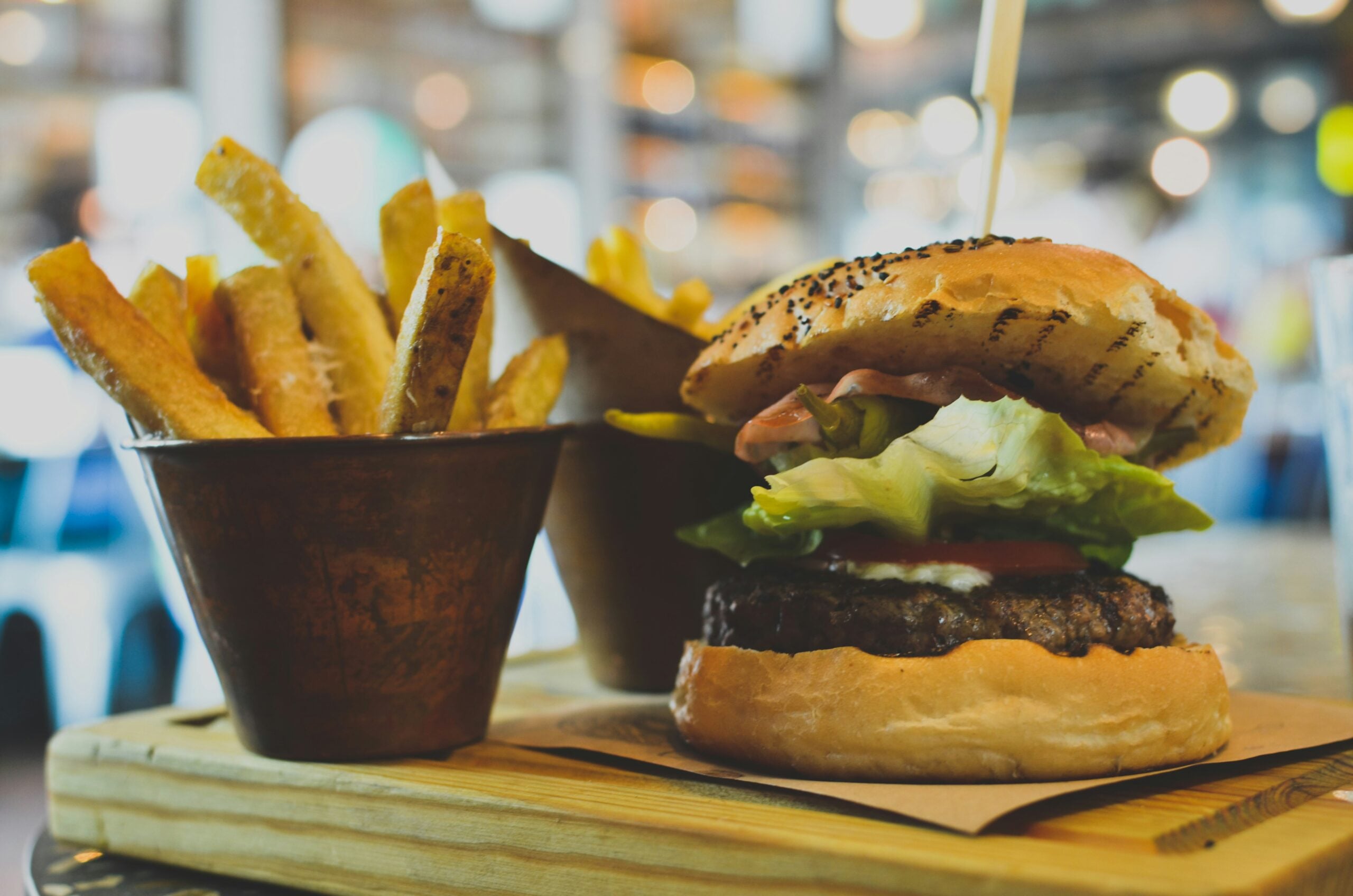 A close-up of a juicy hamburger with lettuce, tomato, and cheese in a bun, next to a metal cup filled with golden French fries, served on a wooden board in a restaurant setting.