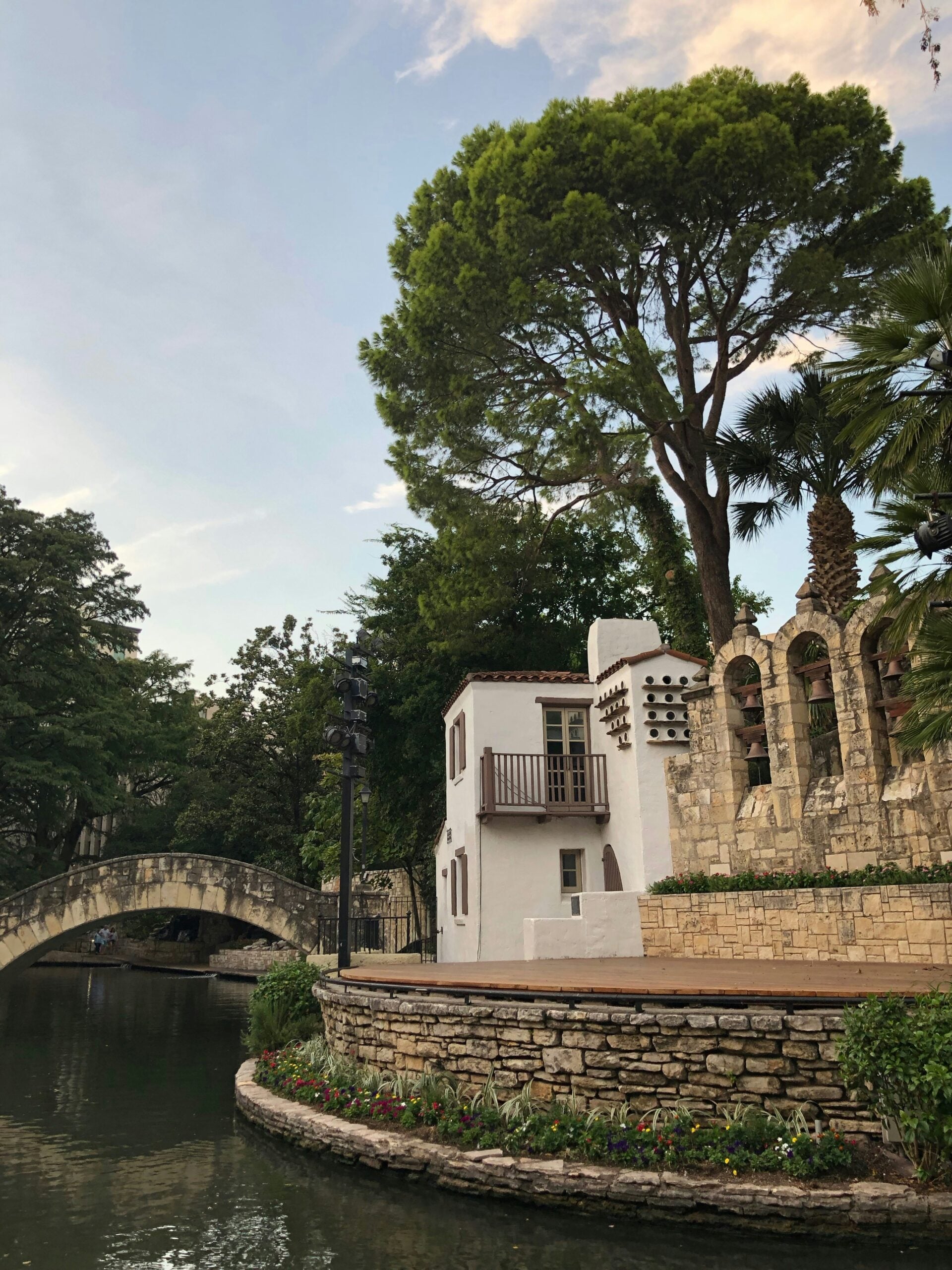 Affordable U.S. vacation spots. A stone building with arched windows and a balcony sits beside a calm river, surrounded by lush trees. A stone footbridge crosses the river in the background, and colorful flowers line the water’s edge.