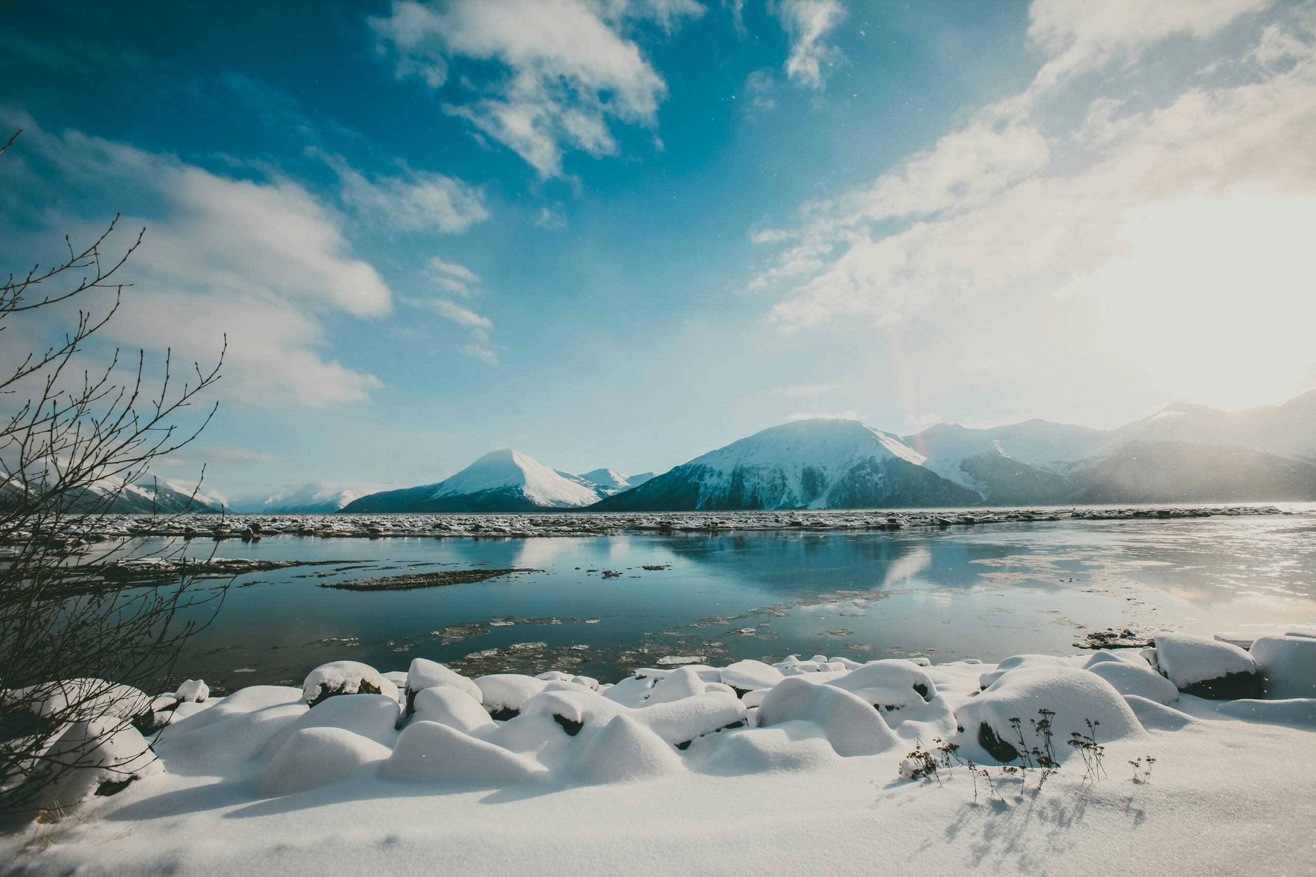 A snowy landscape with a frozen lake, snow-covered rocks in the foreground, and mountains in the distance under a bright blue sky with scattered clouds. The sun is shining from the right side.