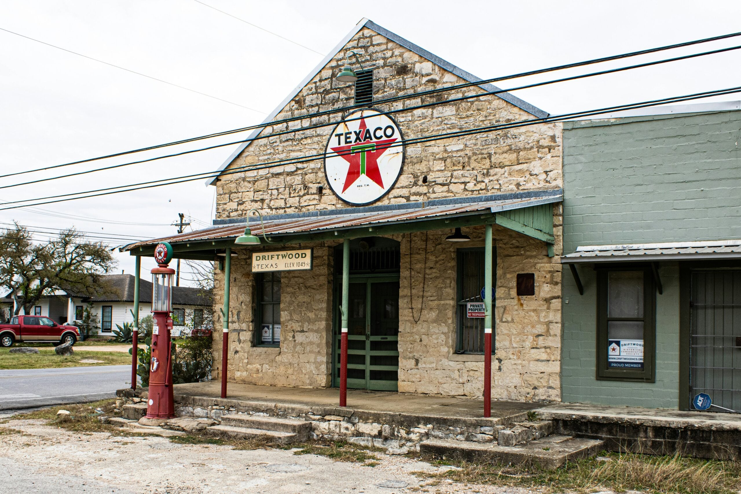 An old stone building with a vintage Texaco sign and red gas pump in front, labeled “Driftwood Texas Store.” A green porch and nearby parked cars suggest a small rural town setting.