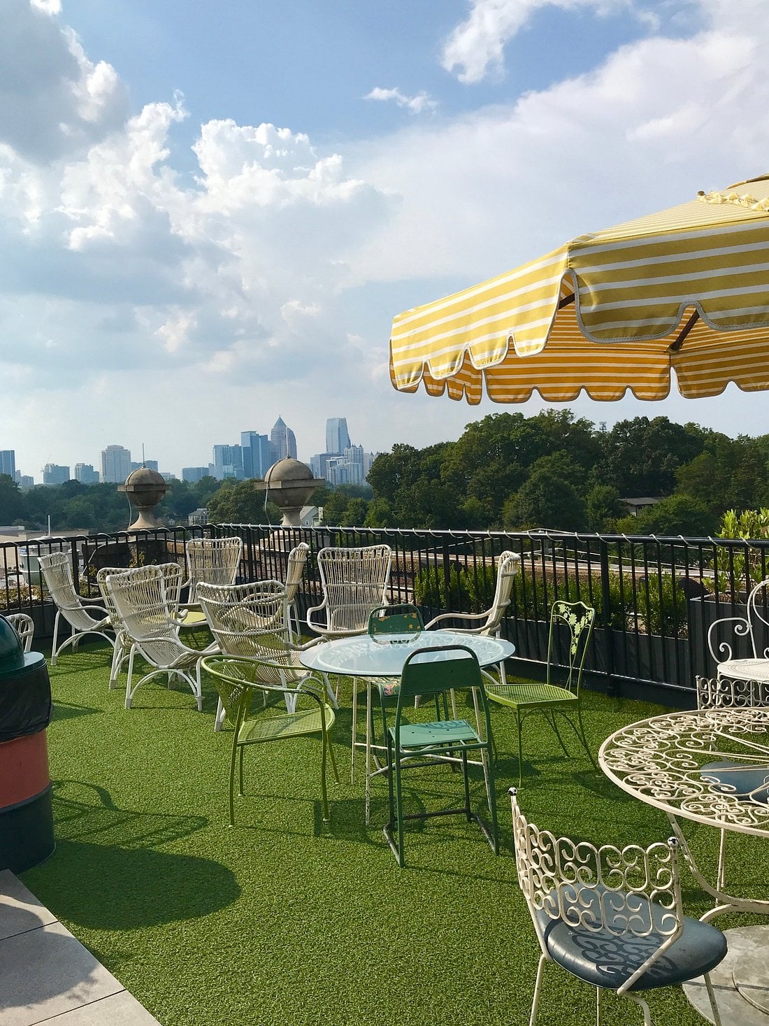 Rooftop patio at a small-town luxury hotel, featuring white metal chairs and tables on artificial grass, a yellow-striped umbrella, and a city skyline visible in the distance under a partly cloudy sky.