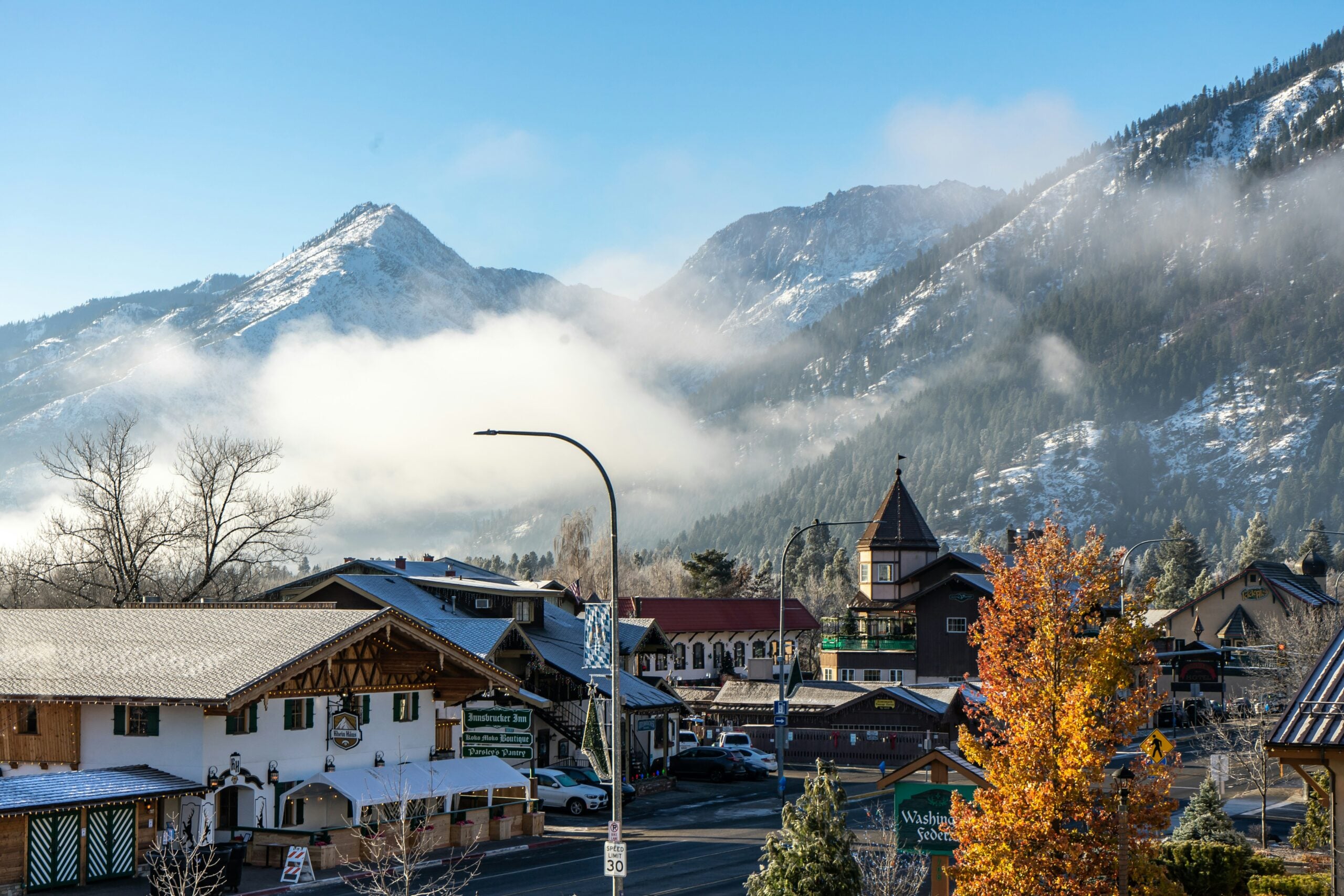 A small mountain town with Bavarian-style buildings, autumn trees, and light snow on rooftops is set against a backdrop of misty mountains under a clear sky.