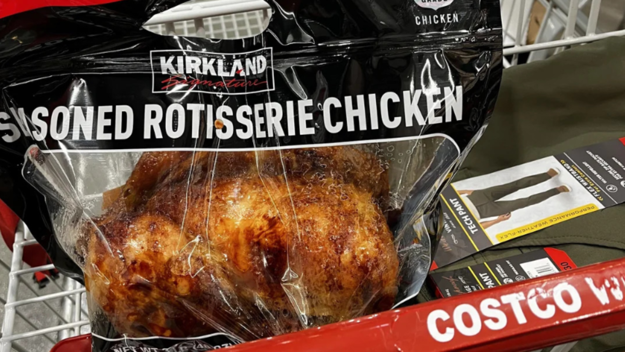 A whole cooked rotisserie chicken in a clear plastic bag sits in a Costco shopping cart next to packaged clothing items. The bag label reads "Seasoned Rotisserie Chicken.