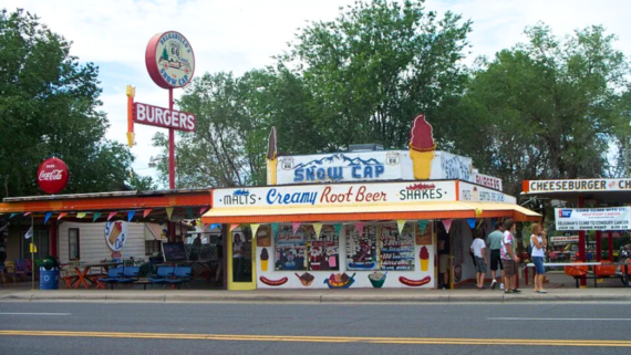 A colorful roadside diner on Route 66 features signs for burgers, malts, root beer, and shakes. People stand out front amid bright decorations and trees, with a classic Route 66 sign displayed proudly on the roof.