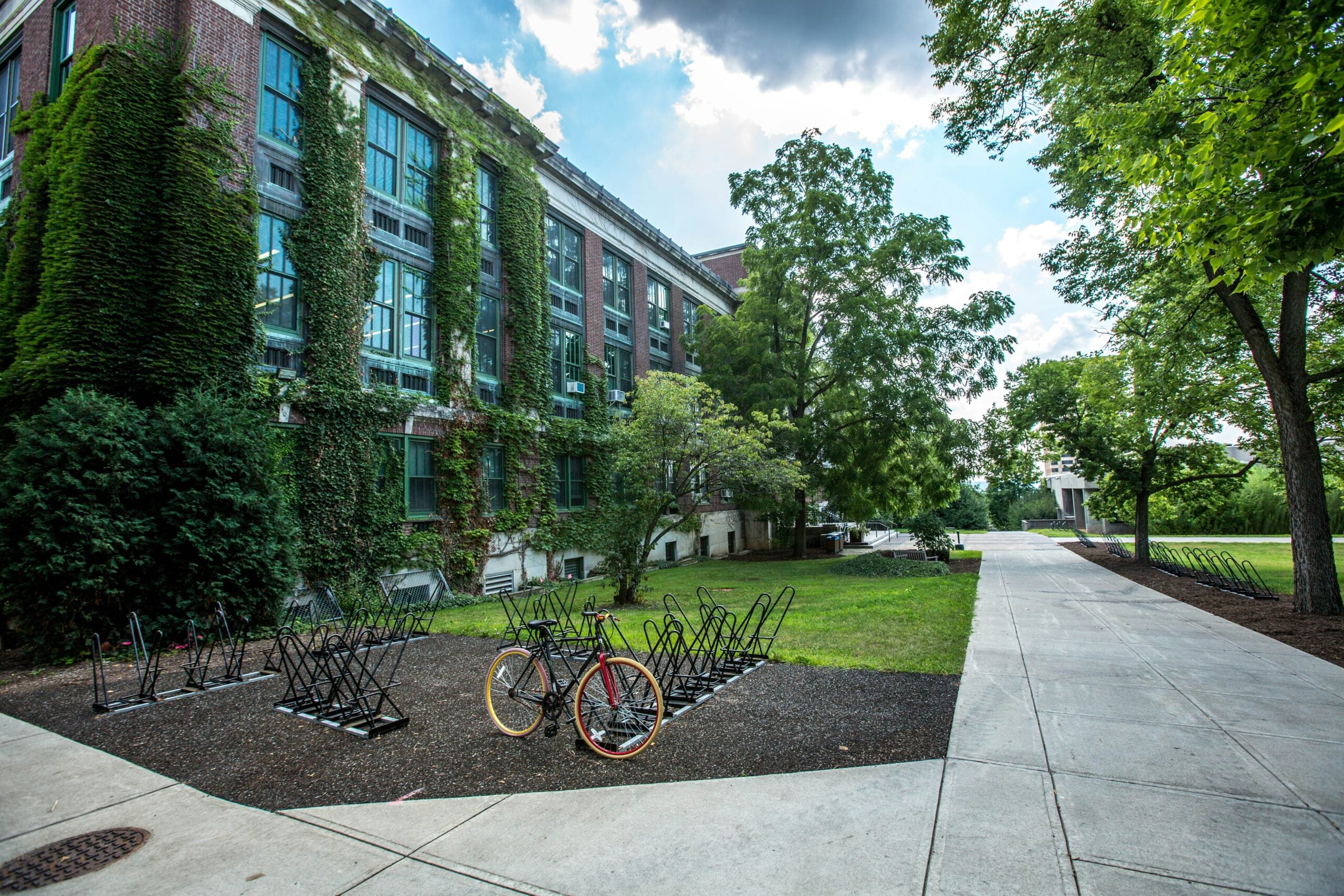 A red bicycle is parked in an outdoor bike rack near a brick building with ivy-covered walls and large windows, surrounded by green trees and a paved walkway under a partly cloudy sky.