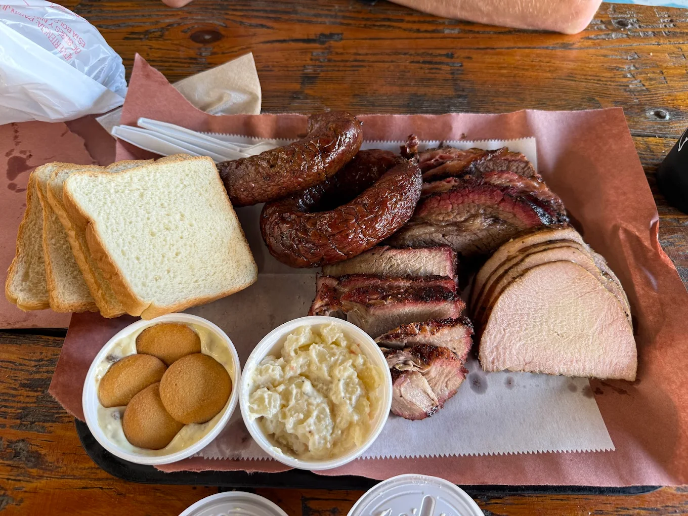 A tray with slices of white bread, sausage links, sliced brisket, sliced pork, banana pudding, and a cup of coleslaw on brown paper atop a wooden table. Plastic utensils and napkin are also visible.
