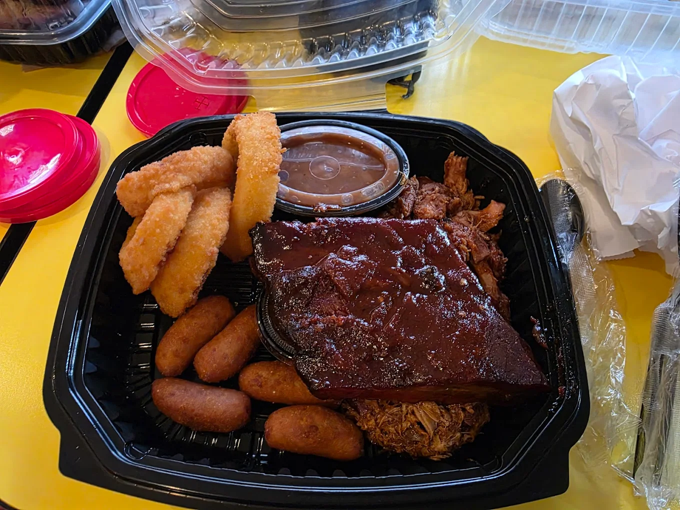 A black takeout container with barbecue ribs, baked beans in a cup, fried okra, hush puppies, and onion rings, set on a yellow table with plastic utensils and takeaway packaging visible.