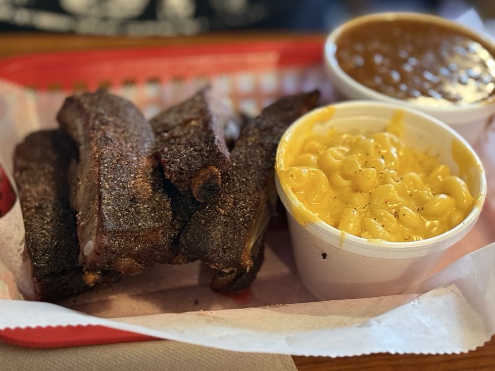 A food tray with several pieces of barbecued ribs, a cup of creamy macaroni and cheese, and a cup of baked beans. The tray is lined with paper.