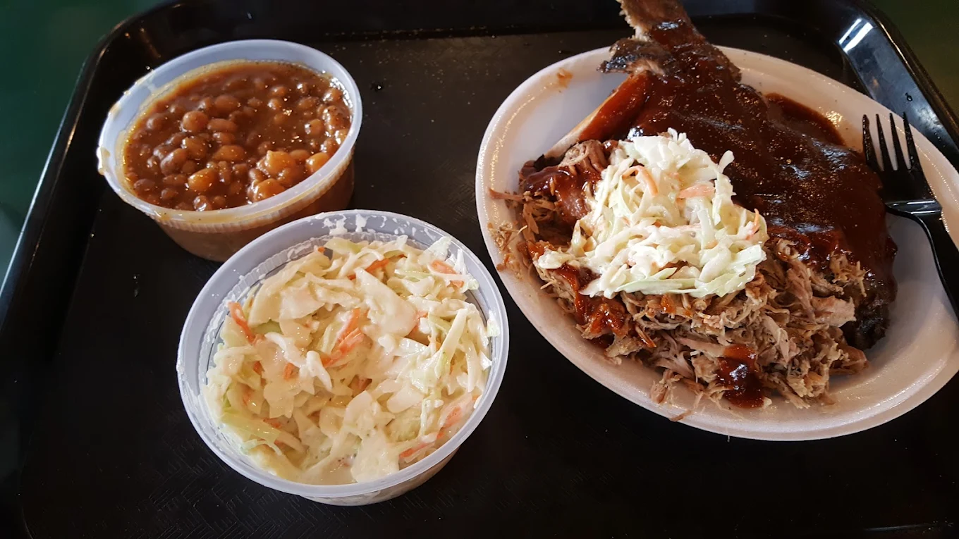 A plate of barbecue pulled pork topped with coleslaw and sauce, served with a side of baked beans in a cup and a separate cup of coleslaw on a black tray.