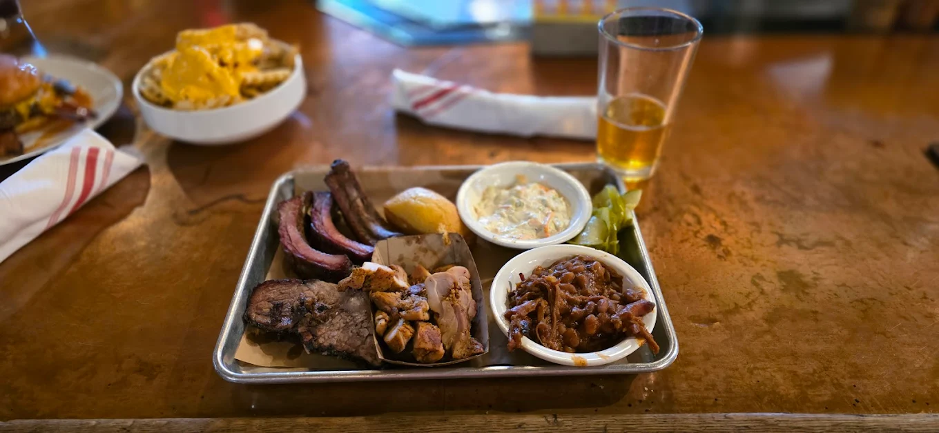 A tray with assorted barbecue meats, coleslaw, baked beans, pickles, and a lemon wedge sits on a wooden table next to a glass of beer and a napkin. Another plate of food is partially visible in the background.