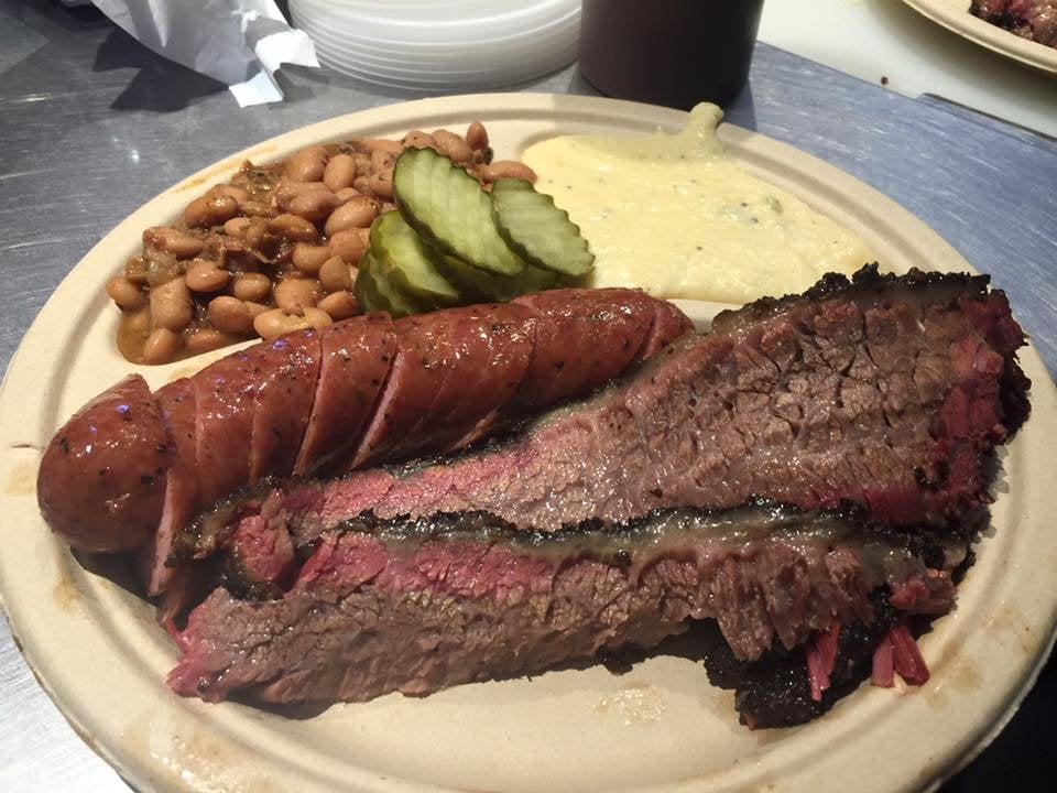 A plate of barbecue with sliced brisket, a sausage, pickles, baked beans, and a serving of creamy mashed potatoes on a tan paper plate.