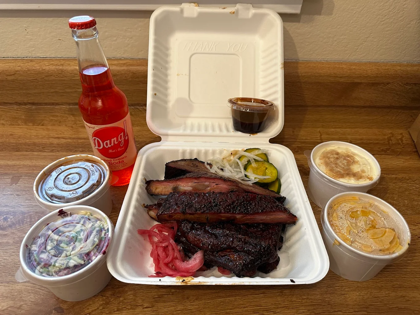 A takeout meal with barbecued ribs and brisket, pickles, onions, and pickled onions in a foam container, surrounded by side dishes, barbecue sauce, and a red Dang! soda bottle on a wooden counter.