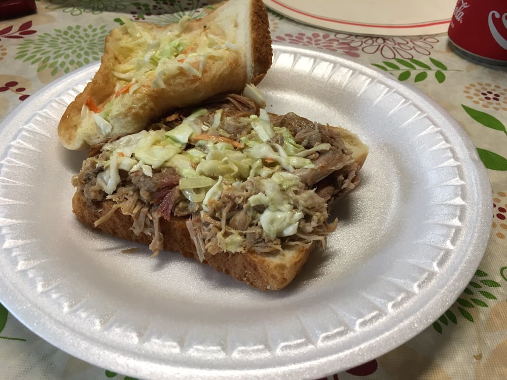 A pulled pork sandwich topped with coleslaw on a white foam plate, resting on a patterned tablecloth—just the kind of comfort you’d find at hole-in-the-wall BBQ spots. A can of soda and another plate are partially visible in the background.