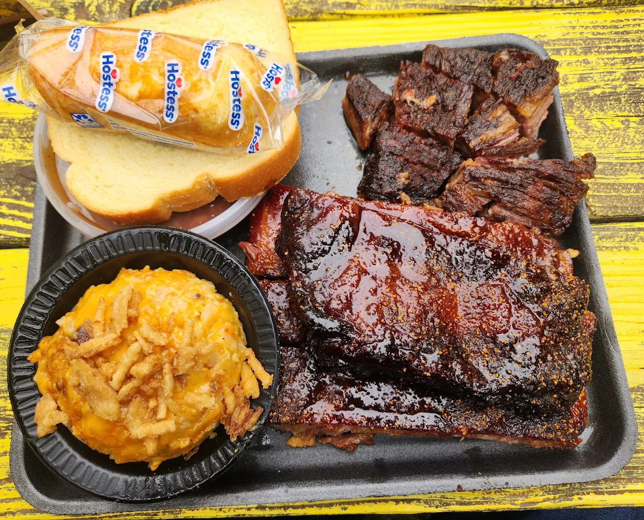 A tray with glazed barbecue ribs, slices of smoked brisket, cheesy potato casserole, white bread, and a packaged Hostess dessert on a yellow wooden table.