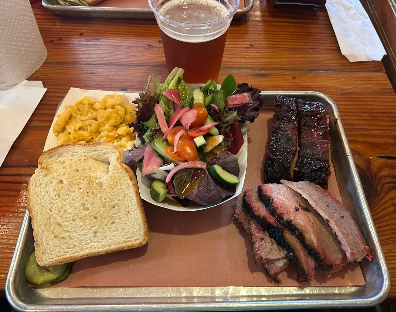 A tray reminiscent of classic hole-in-the-wall BBQ spots: sliced brisket, ribs, macaroni and cheese, white bread, a fresh side salad, pickle slices, and a plastic cup of beer—all served on a rustic wooden table.