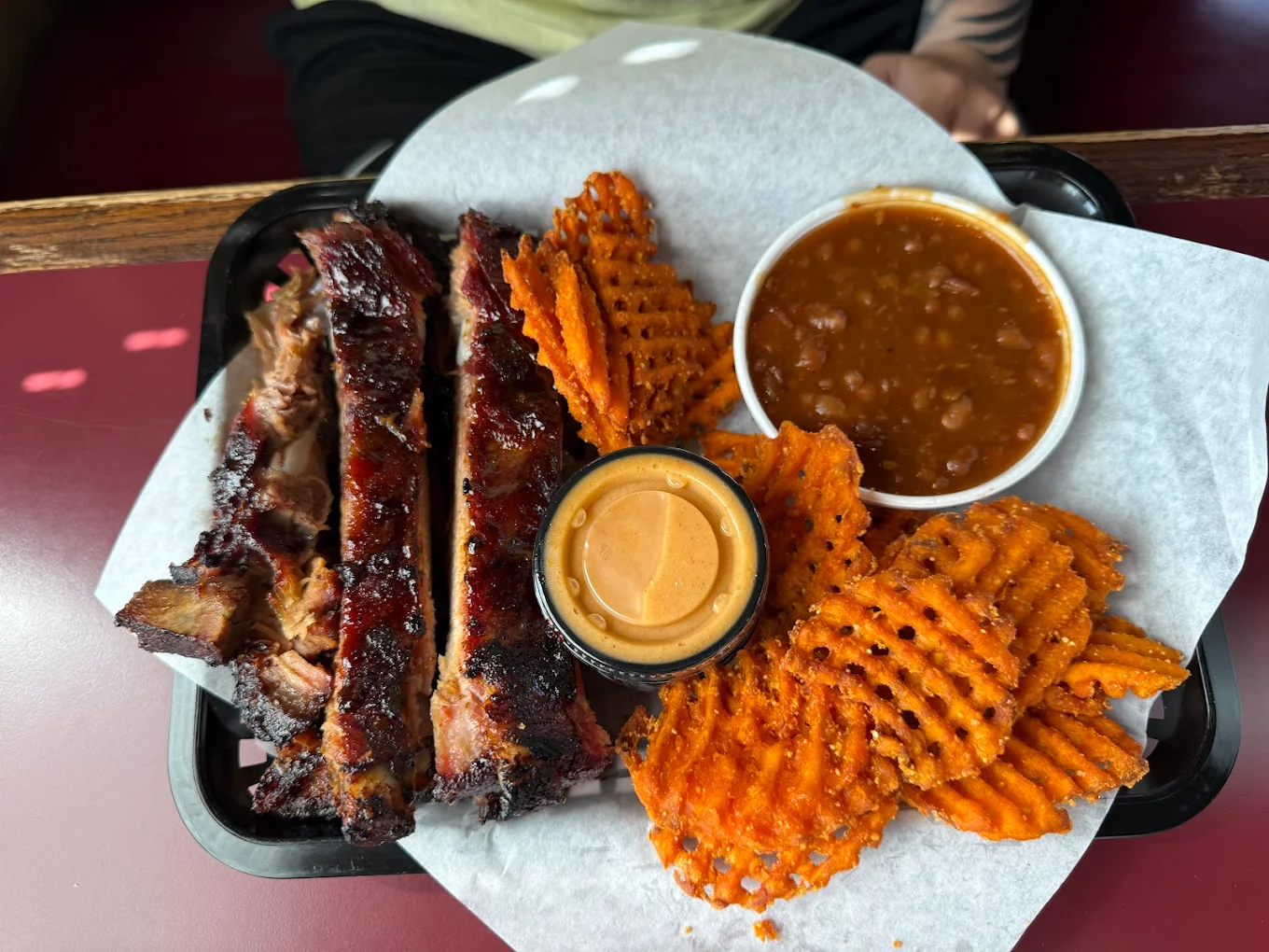 A tray with barbecue ribs, a small cup of dipping sauce, sweet potato waffle fries, and baked beans—just the kind of hearty fare you’d find at the best hole-in-the-wall BBQ spots, all served on parchment paper.