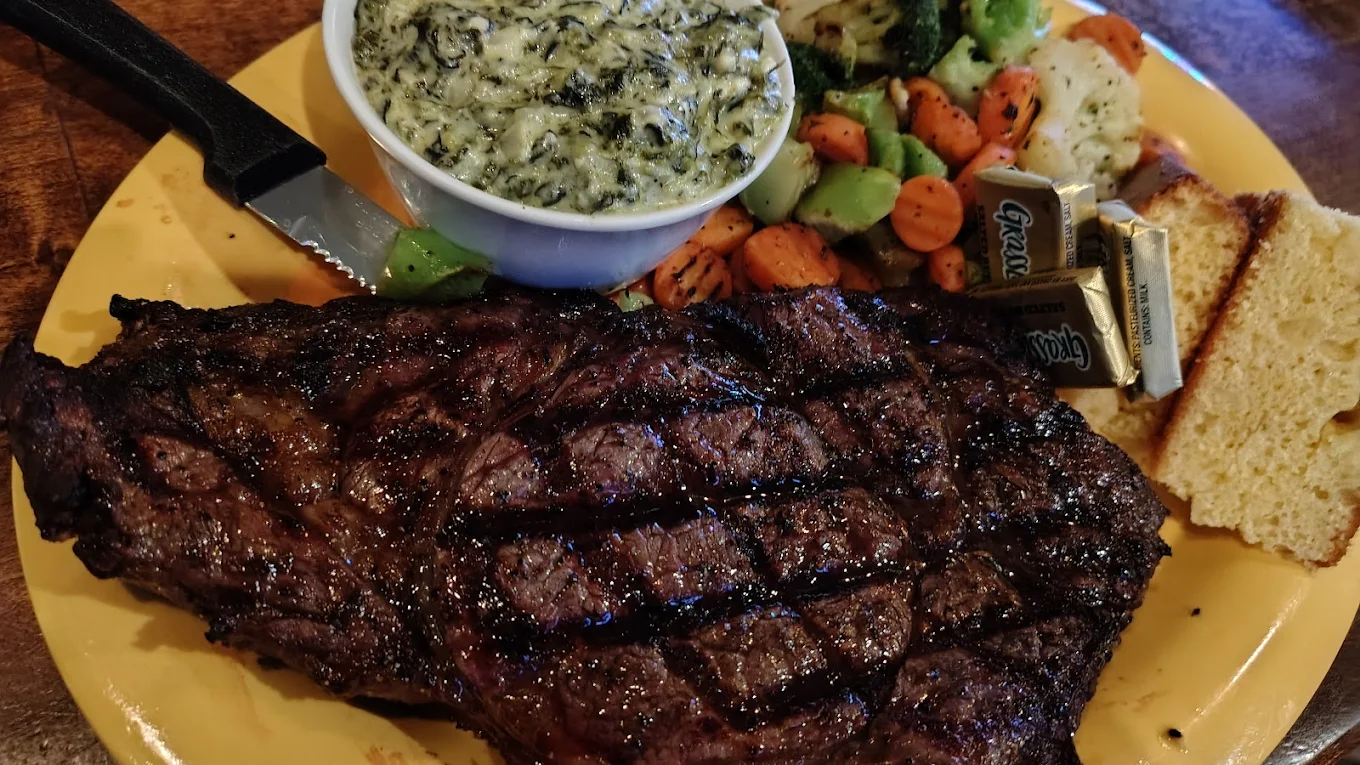 A grilled steak with char marks is served with creamy spinach, sautéed vegetables, cornbread, and butter on a yellow plate—just the kind of hearty meal you’d find at hole-in-the-wall BBQ spots.