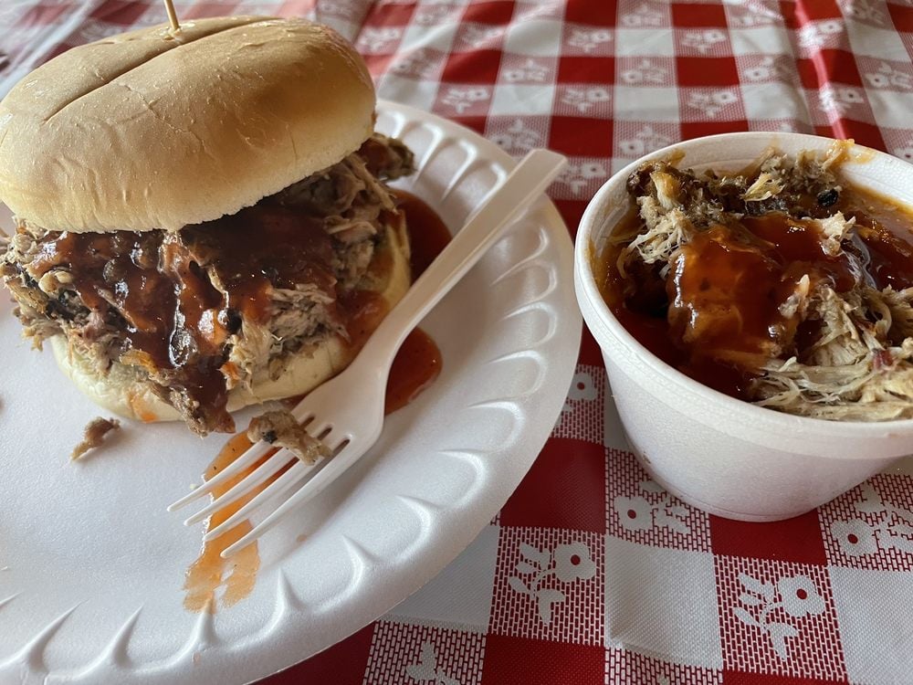 A pulled pork sandwich with barbecue sauce on a white bun sits on a paper plate next to a cup filled with more sauced pulled pork, on a red-and-white checkered tablecloth. A plastic fork is on the plate.