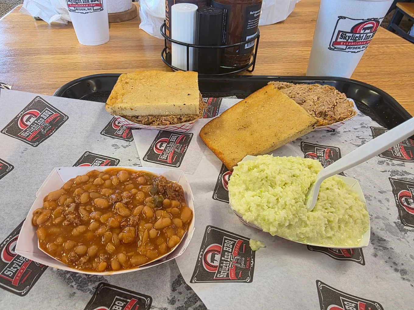 A tray with baked beans, coleslaw, pulled pork, and two pieces of cornbread on BBQ-branded paper, along with two large drinks in white cups on a wooden table.
