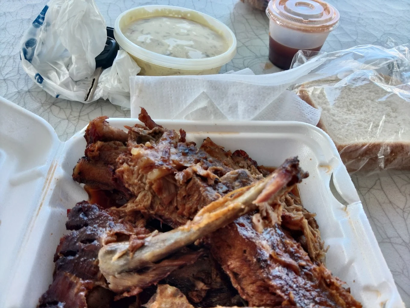 A takeout container filled with barbecue ribs and pulled meat sits on a table, next to slices of white bread, a container of sauce, and a tub of creamy soup or mashed potatoes.