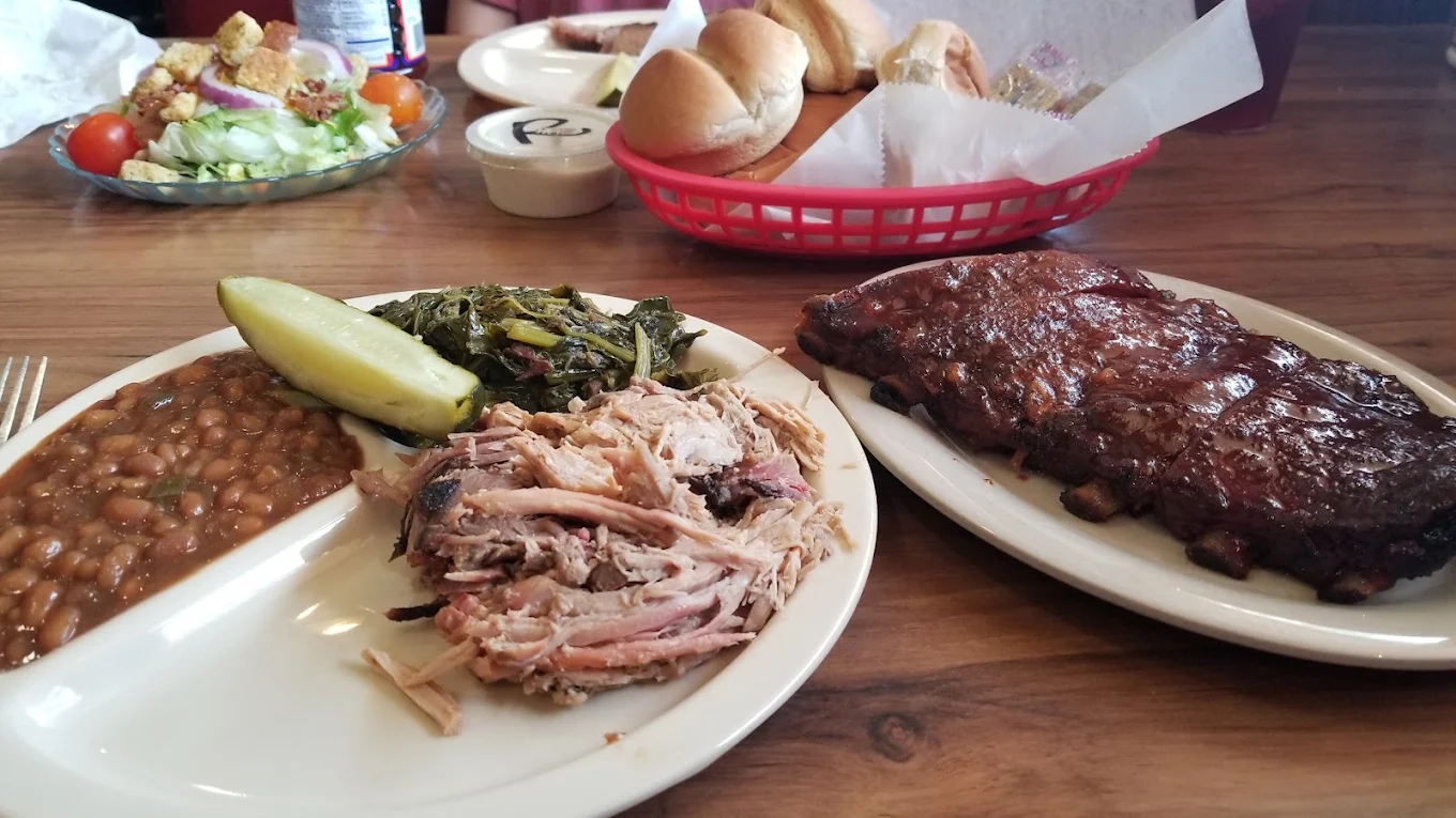 A table with a plate of baked beans, pulled pork, collard greens, and a pickle; a plate of barbecue ribs; a salad; and a basket of bread rolls in the background.