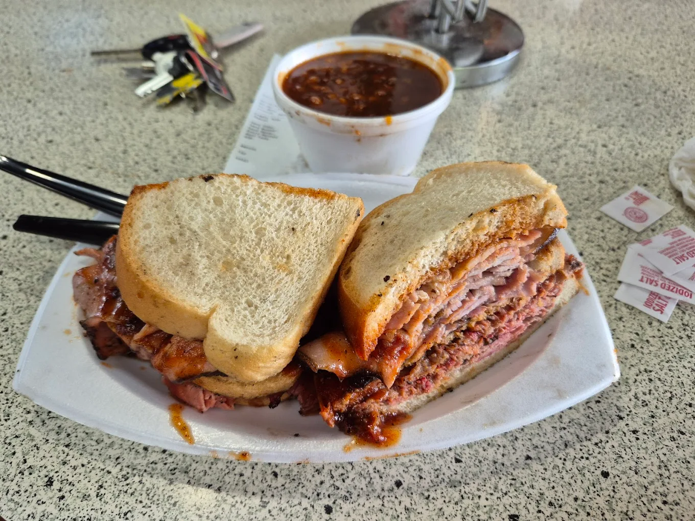 A plate with a thick barbecue meat sandwich on white bread, served with a cup of baked beans, plastic utensils, and packets of salt on a speckled countertop, with keys and a receipt in the background.