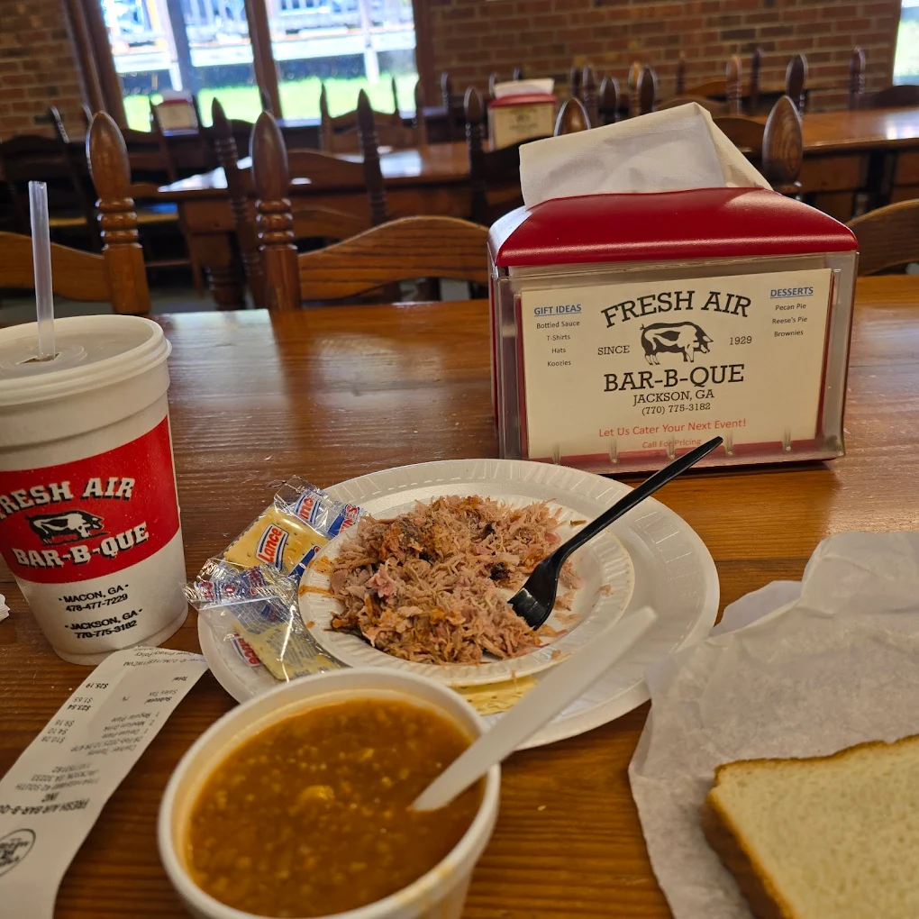 A plate of pulled pork with a fork, baked beans, white bread, saltines, a drink cup, and a receipt on a wooden table at Fresh Air Bar-B-Que restaurant, with tables and chairs in the background.