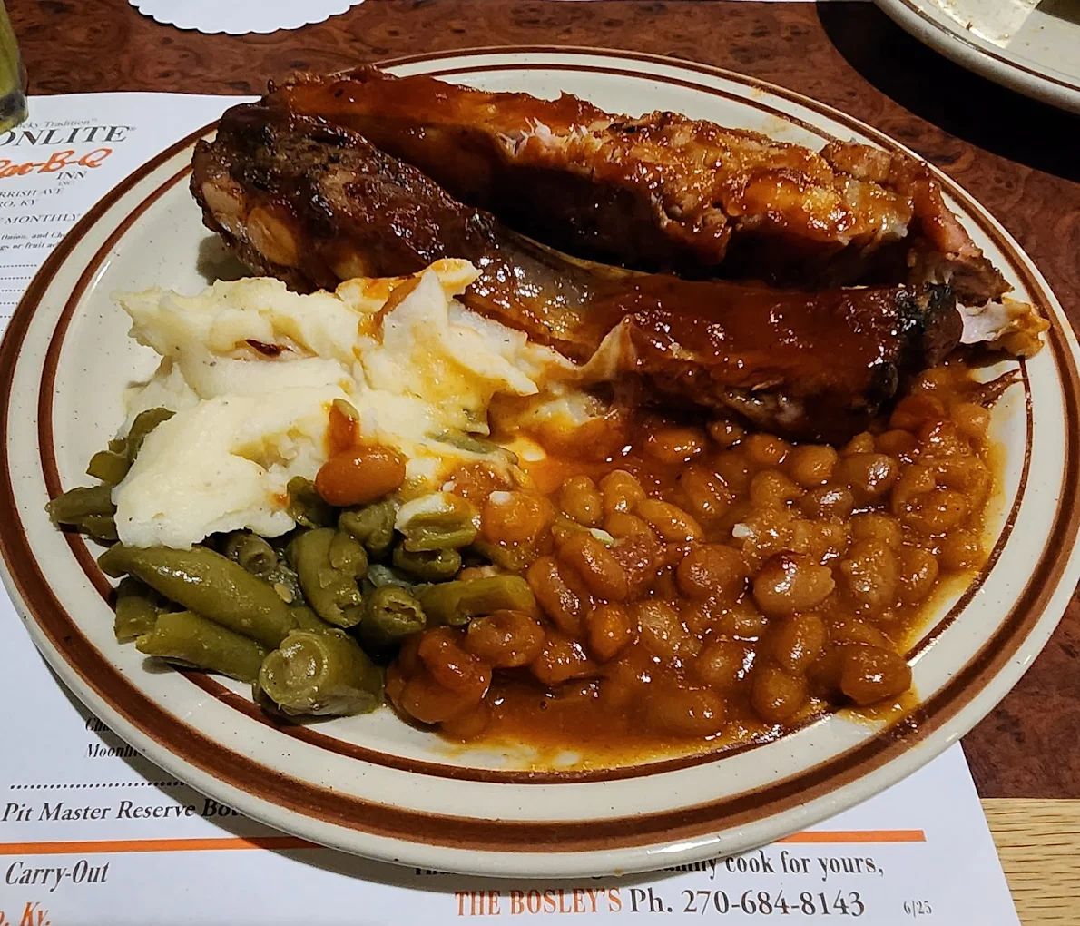 A dinner plate with saucy barbecued ribs, mashed potatoes with gravy, baked beans, and cooked green beans on a restaurant table.