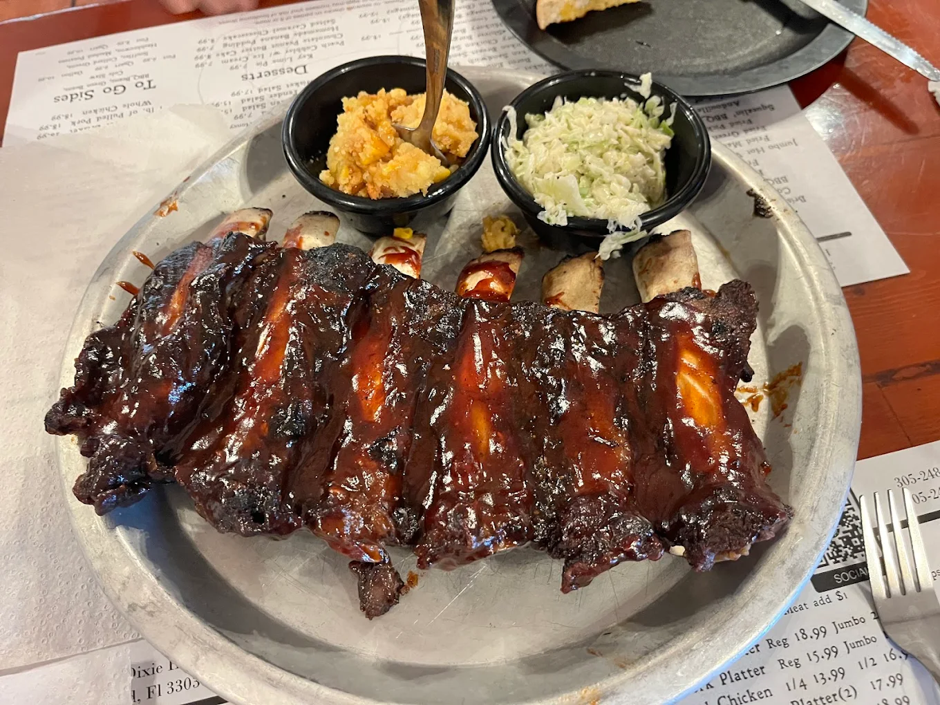 Hole-in-the-wall BBQ spots. A plate of glazed barbecue ribs served with two small bowls, one containing coleslaw and the other containing a cornbread dish, on a table in a restaurant.