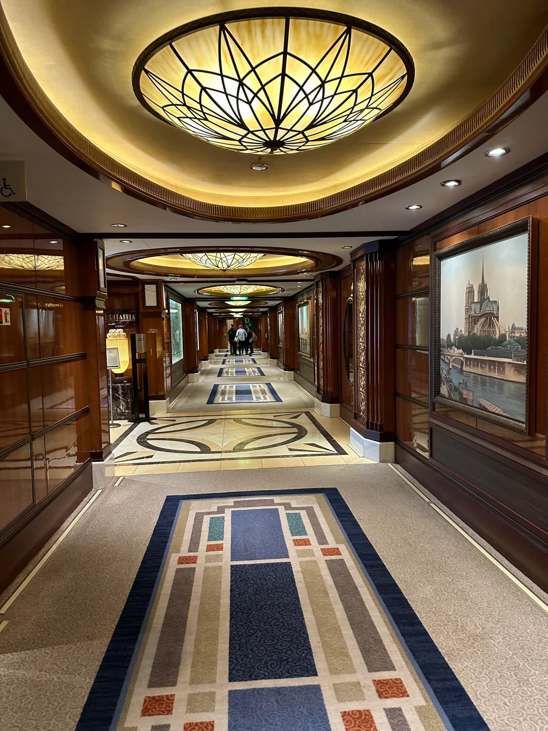 Elegant hallway with art deco lighting, patterned carpets, and wood-paneled walls—just what you’d expect from the Best Cruise Line 2026. Glass display cases and framed artwork line the corridor leading to a well-lit area where guests gather.