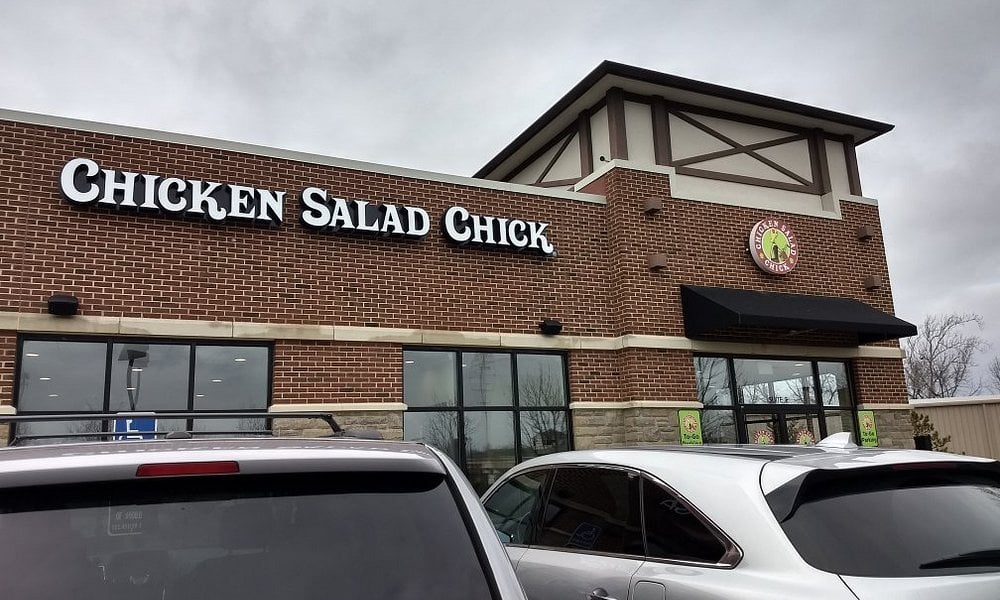 Exterior of a Chicken Salad Chick restaurant with brick walls, black awnings, and parked cars in front. The sky is overcast, and the building’s sign is clearly visible above the entrance.