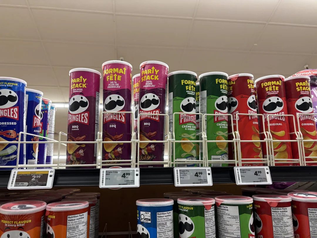Cans of Pringles chips in various flavors, including BBQ and Original, are displayed on a supermarket shelf. The cans are labeled "Party Stack" and "Format Fête" with price tags below them.