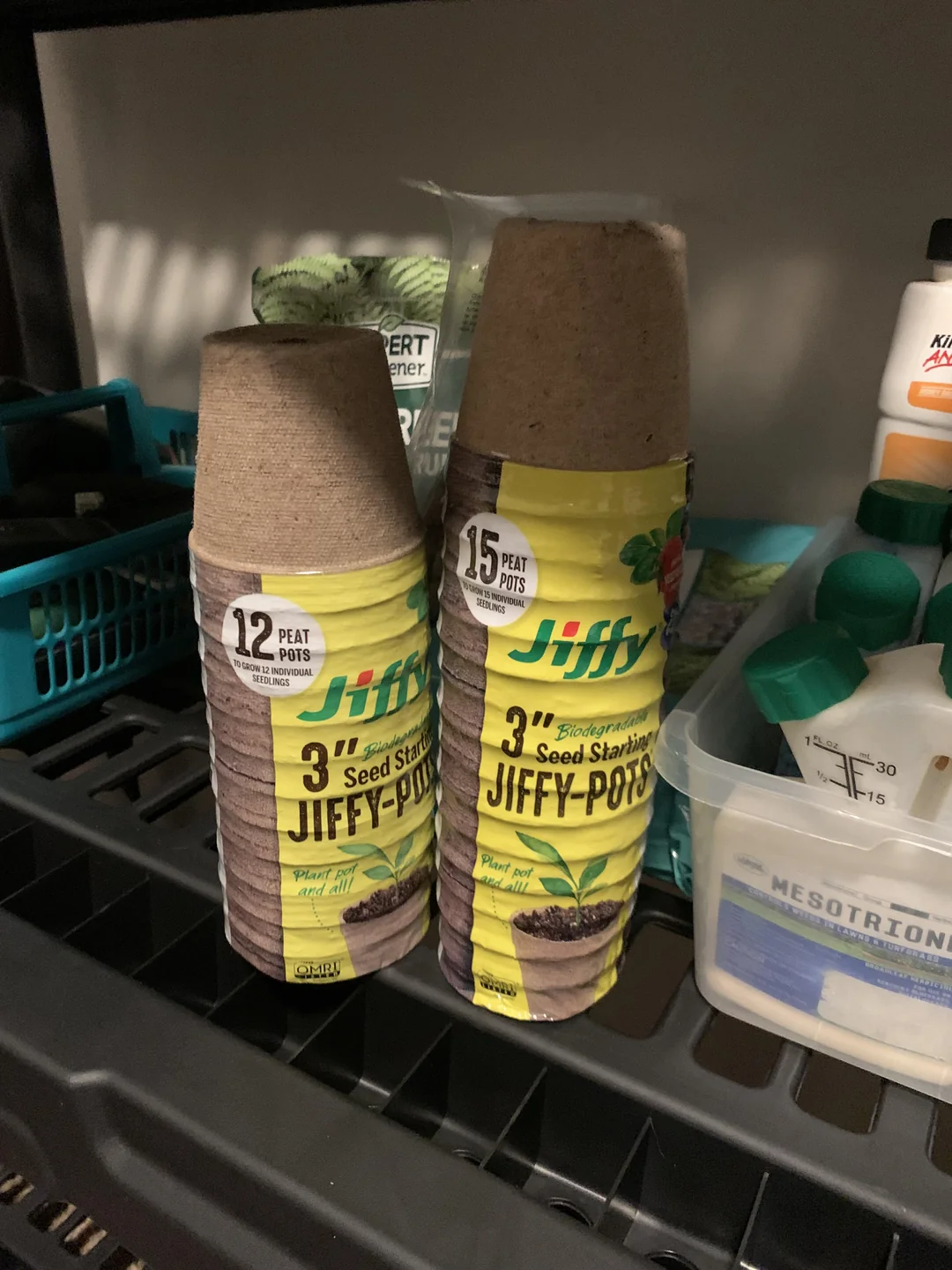 Two stacks of Jiffy 3-inch seed starting peat pots, one pack of 12 pots and one pack of 15 pots, sit on a shelf with other gardening supplies in the background.