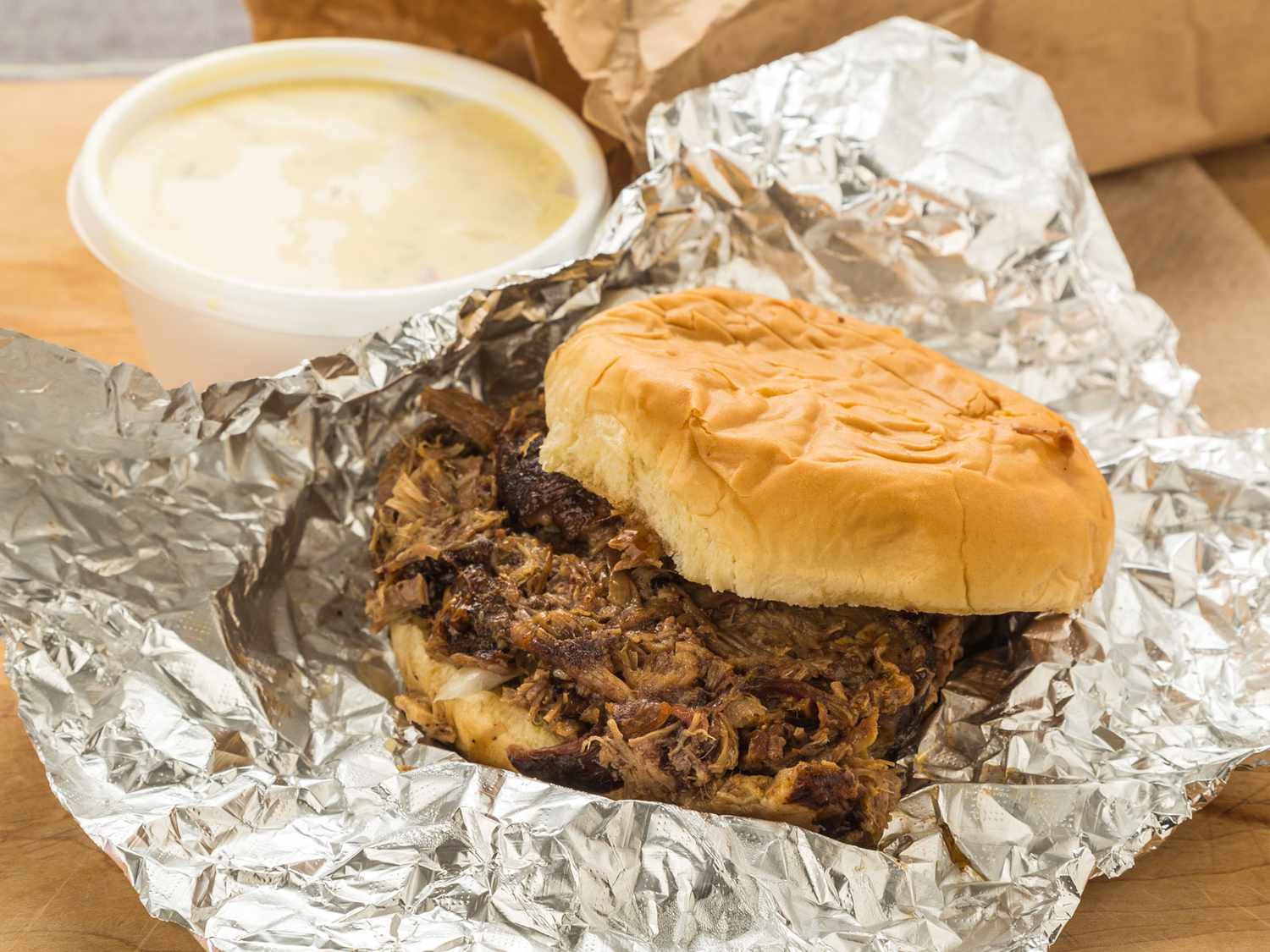 A pulled pork sandwich on a soft bun, wrapped in crinkled aluminum foil, sits next to a white takeout cup of creamy soup on a wooden surface.