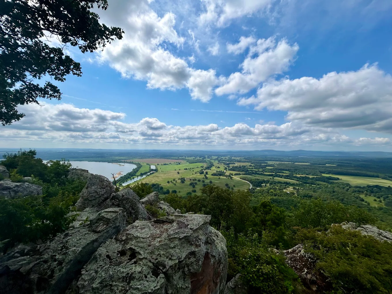 View from a rocky hillside overlooking a river, green fields, and forests under a blue sky with scattered clouds. The landscape stretches into the distance, framed by tree branches on the left.