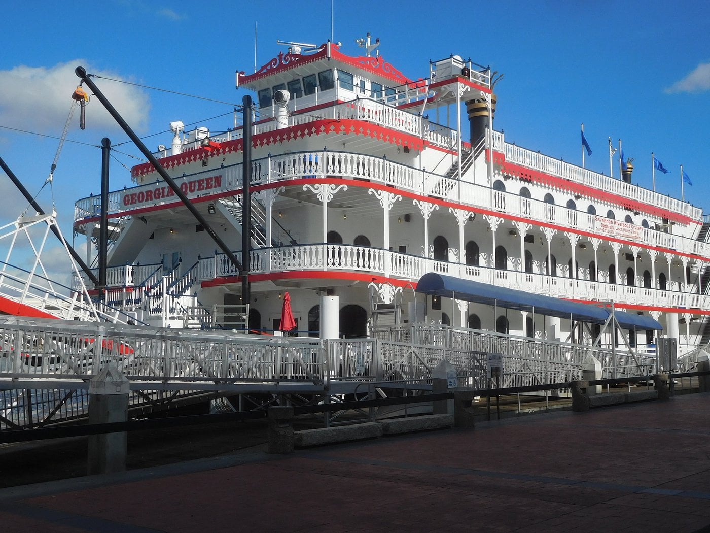 A large white and red riverboat named "Georgia Queen" is docked at a pier, featuring multiple decks with ornate railings and flags, under a partly cloudy blue sky.