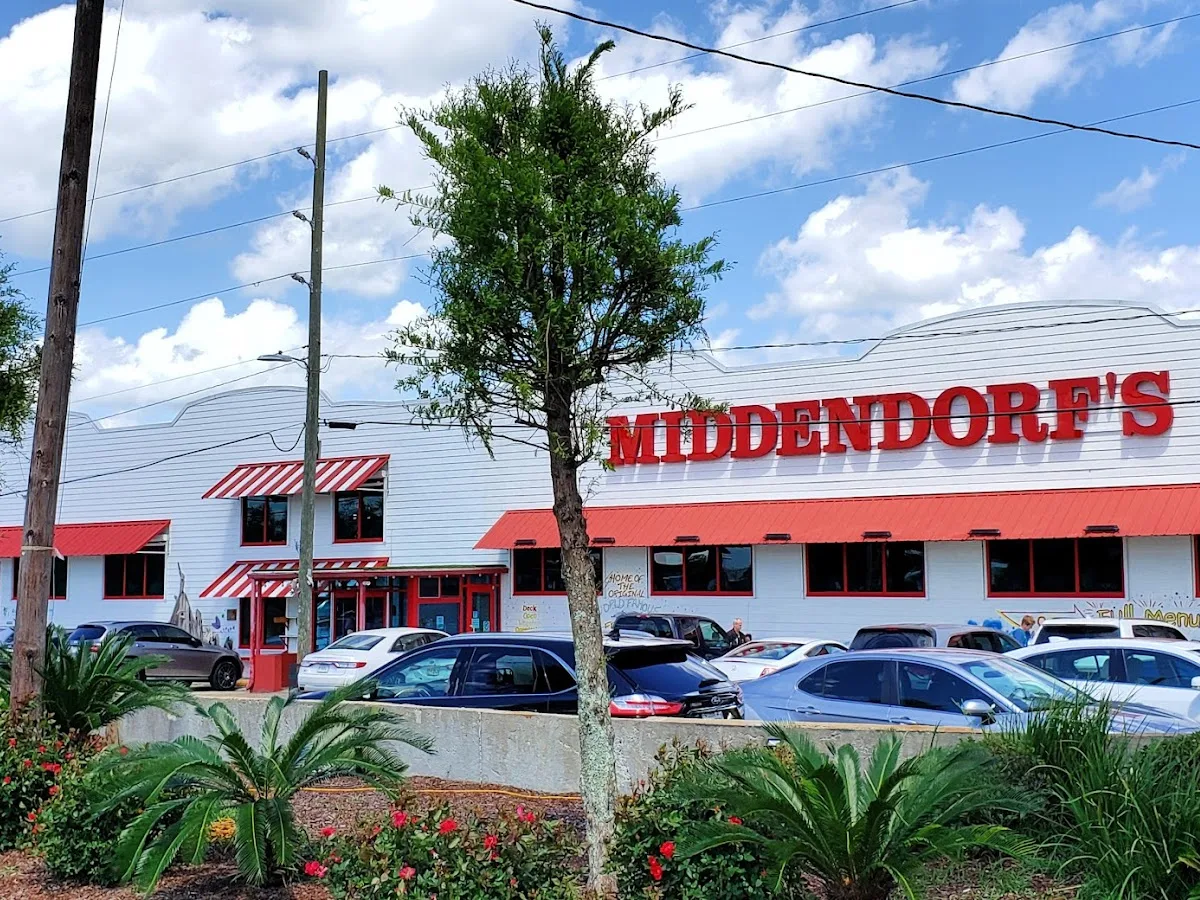 A white building with red trim, large red letters spelling "Middendorf's" on the front, several parked cars, palm plants, and a tree in the foreground under a partly cloudy sky.