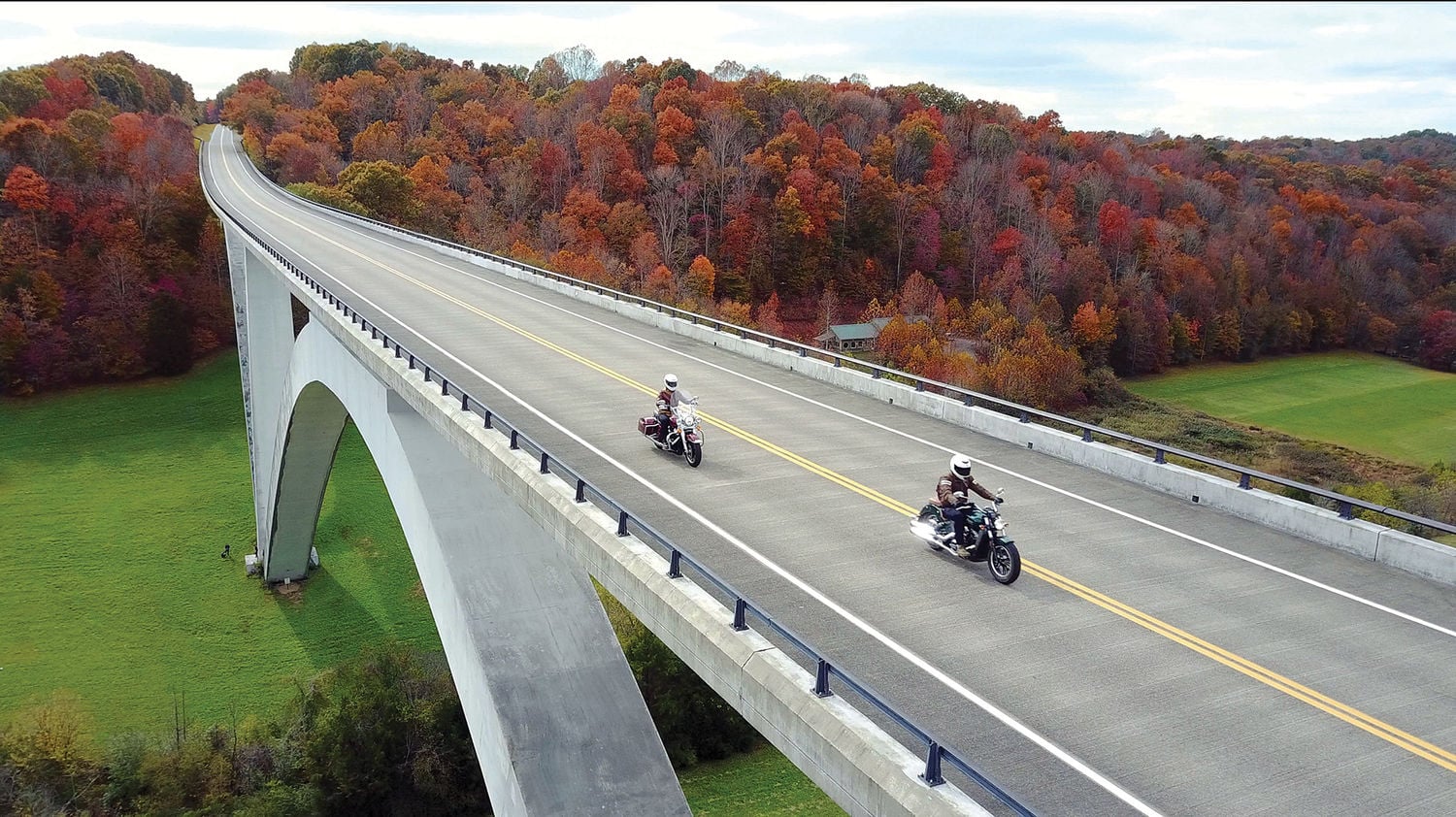 Two people ride motorcycles across a tall, curved bridge surrounded by autumn trees with colorful foliage. The bridge stands over green fields and leads into a forested area under a partly cloudy sky.