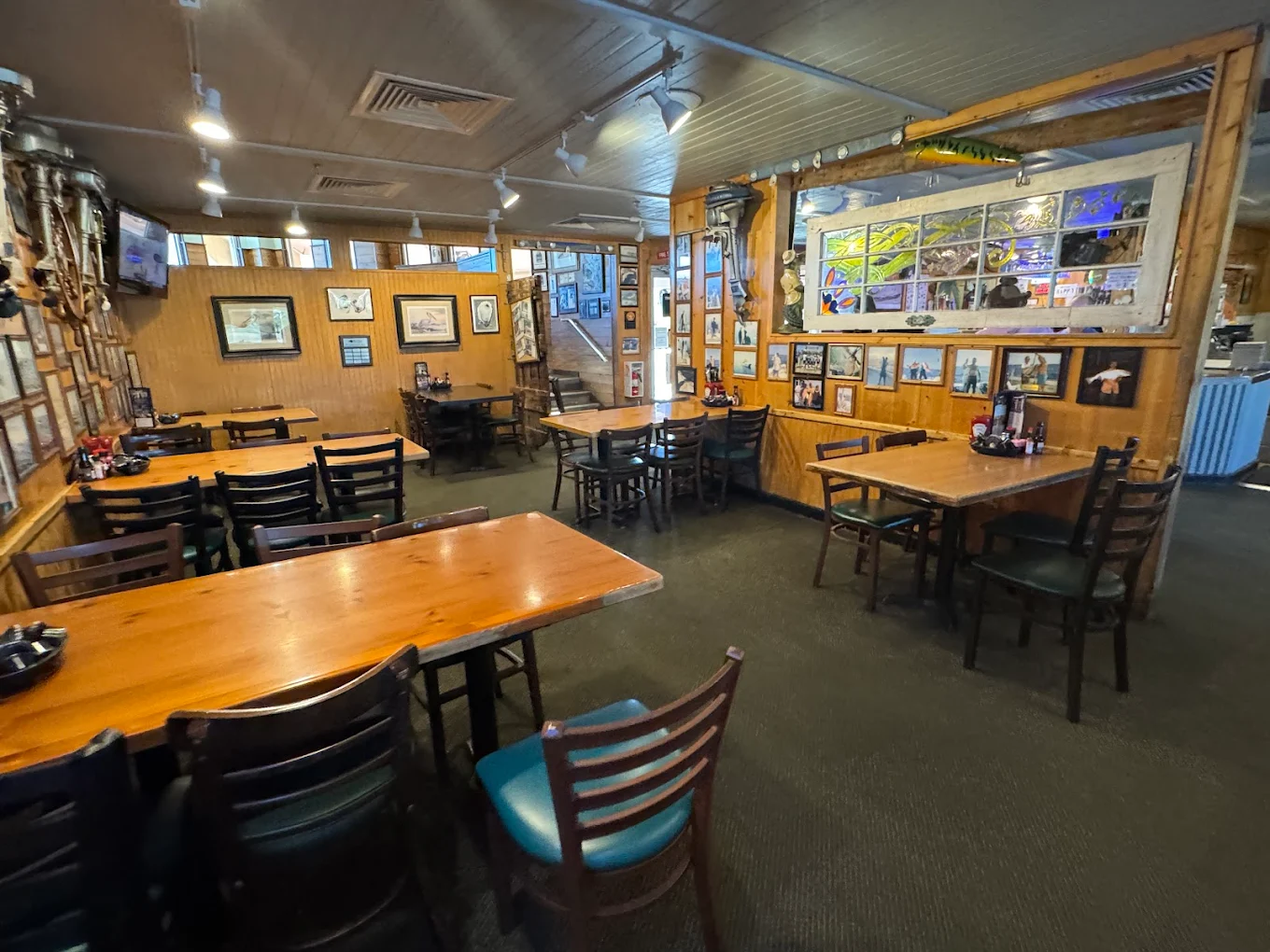A cozy restaurant interior with wooden tables and chairs, walls decorated with various framed photos and memorabilia, and a window with glass panes. The atmosphere is casual and inviting, with soft lighting.