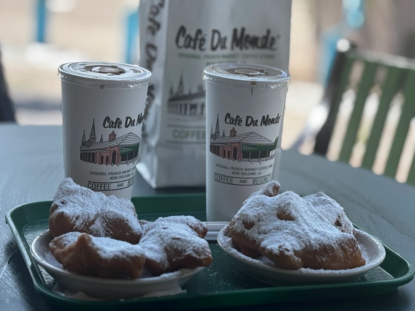 Two plates of powdered sugar-covered beignets and two takeaway coffee cups labeled "Café Du Monde" sit on a green tray, with a Café Du Monde paper bag and outdoor seating visible in the background.