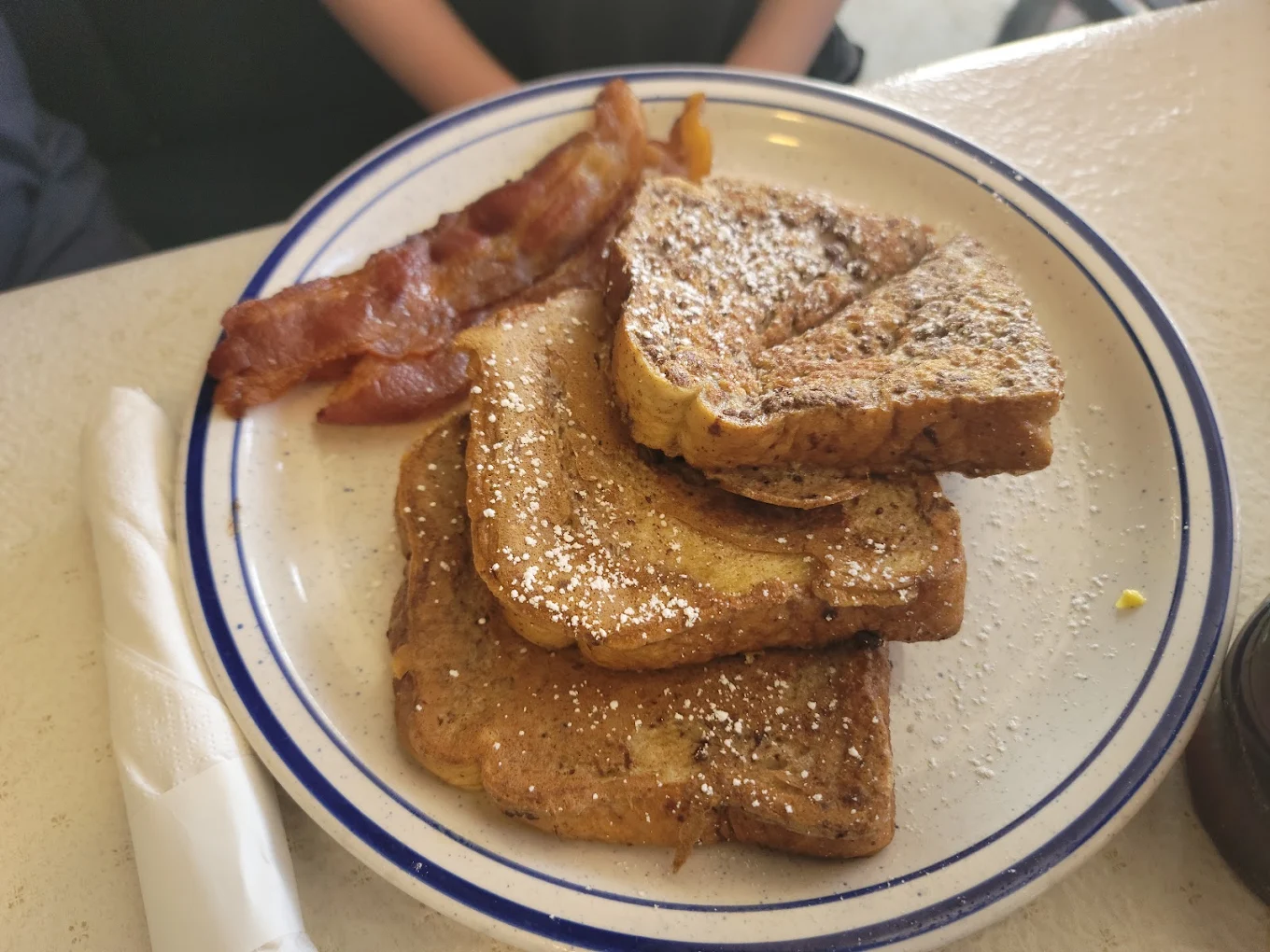 A plate with three slices of French toast topped with powdered sugar, served with several strips of crispy bacon. A napkin and part of a drink are visible nearby.