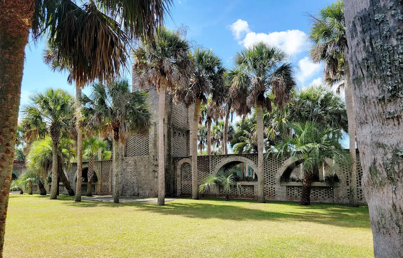 A historic-looking stone building with arched doorways and windows stands among tall palm trees on a grassy lawn under a bright blue sky with scattered clouds.